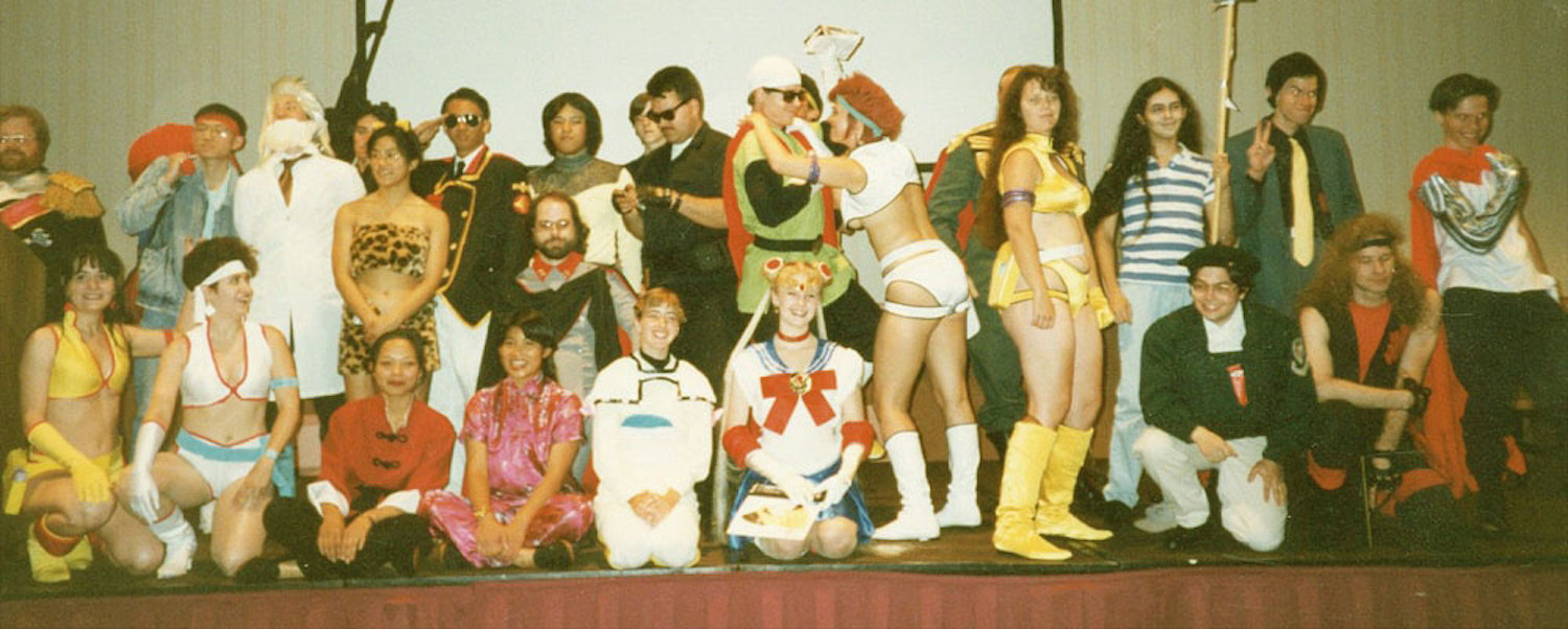 A group of cosplayers onstage at a convention during the 80s. 