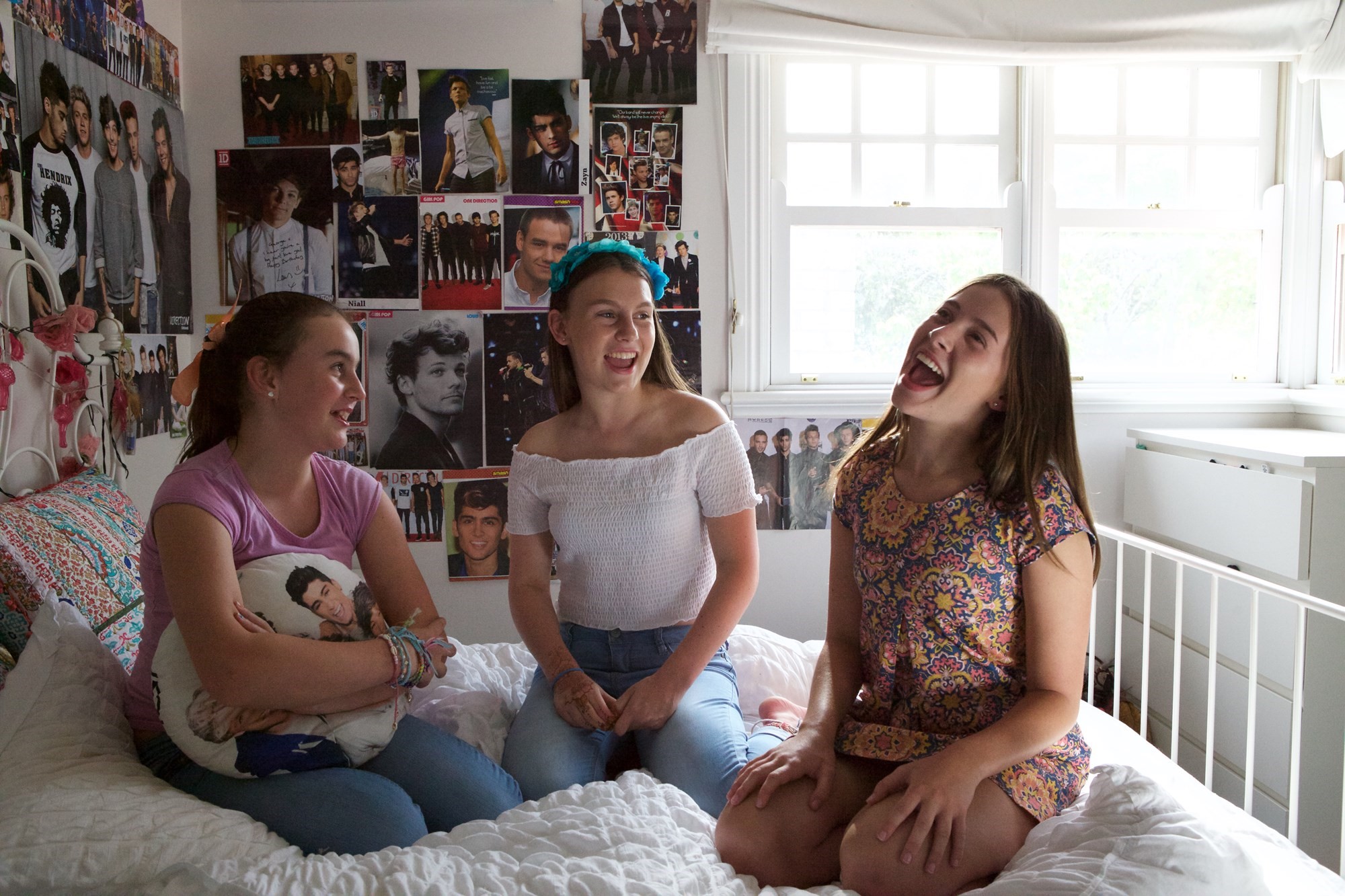 Three girls sitting on a bed surrounded by One Direction paraphernalia. 