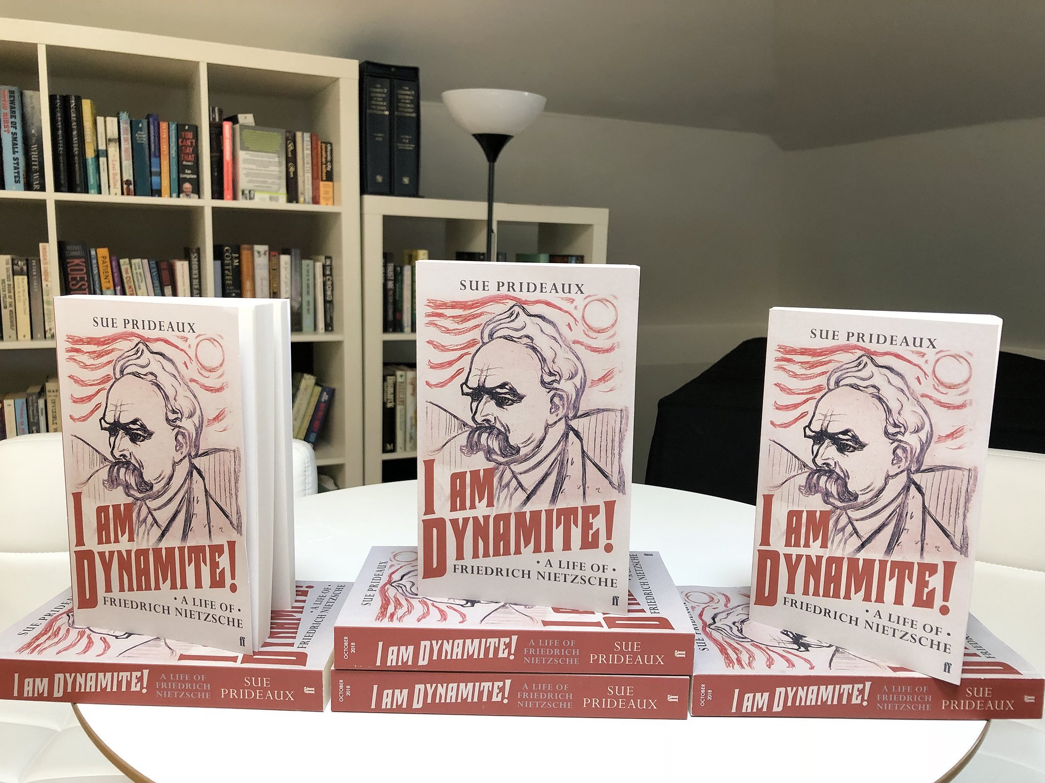 On a white table, several copies of Sue Prideaux's book, "I Am Dynamite! A life of Fredrich Nietzsche" are displayed. Three copies are stood upright on others that have been laid on their sides. The books themselves are white, featuring a sketch of Nietzsche in black with red accents on the front cover. The title is formatted in large, bold, red lettering.