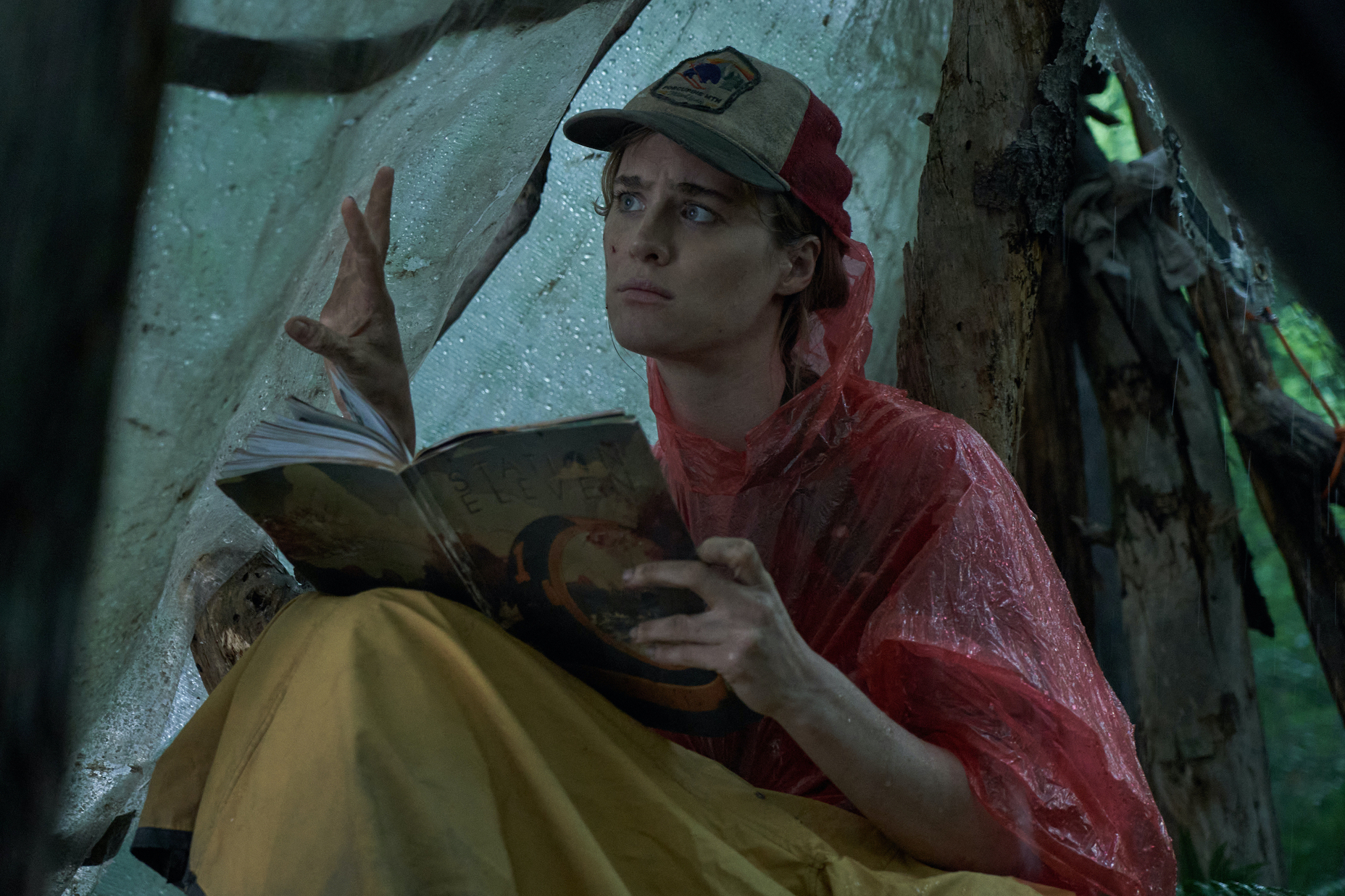 Kirsten (Mackenzie Davis) sits in a tent while reading the book Station Eleven. She is holding one hand up to the wall of the tent, and she looks concerned.