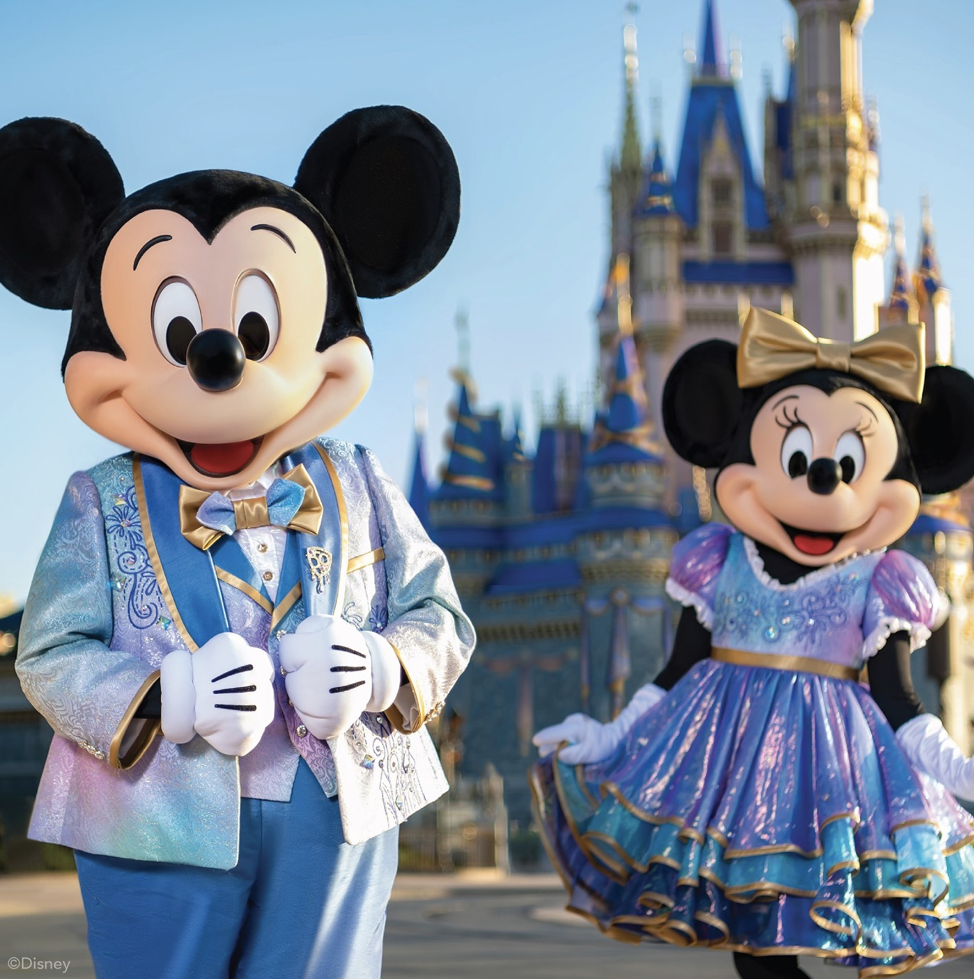 Mickey and Minnie Mouse pose in front of Cinderella's Castle in Magic Kingdom of Walt Disney World, Orlando, Florida. They are dressed in special outfits to celebrate the 50th anniversary of the theme park. Their clothes are a tie-dye of pastel blue and pink with golden accents.