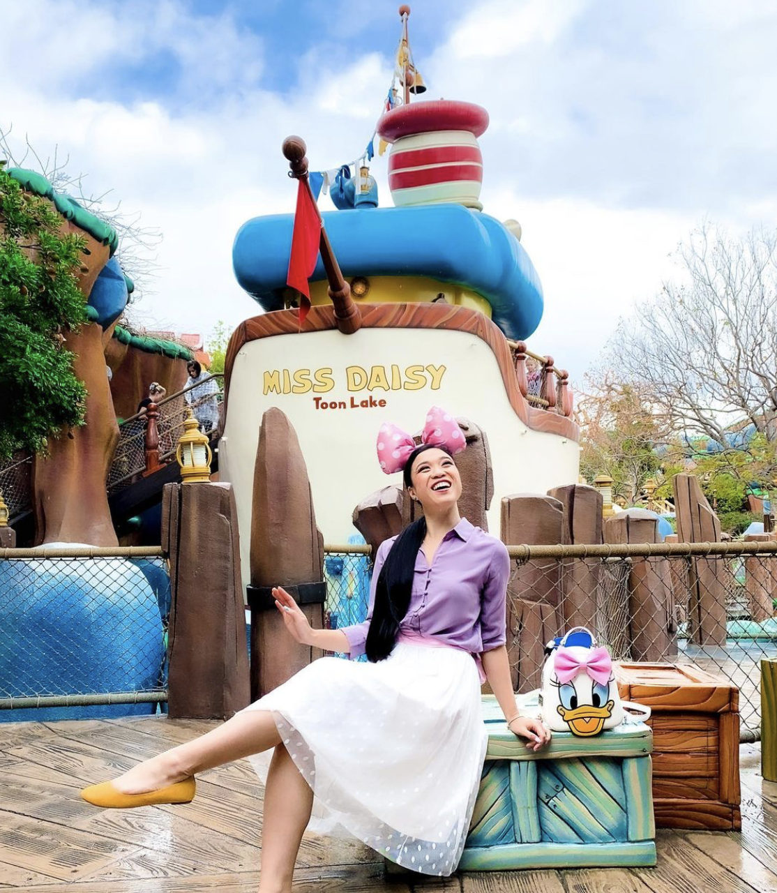 A woman dressed in clothes that resemble Daisy Duck sits and poses in front of a fake boat at a Disney theme park. A small backpack with Daisy's face on it sits beside her. The boat reads, "Miss Daisy Toon Lake." 