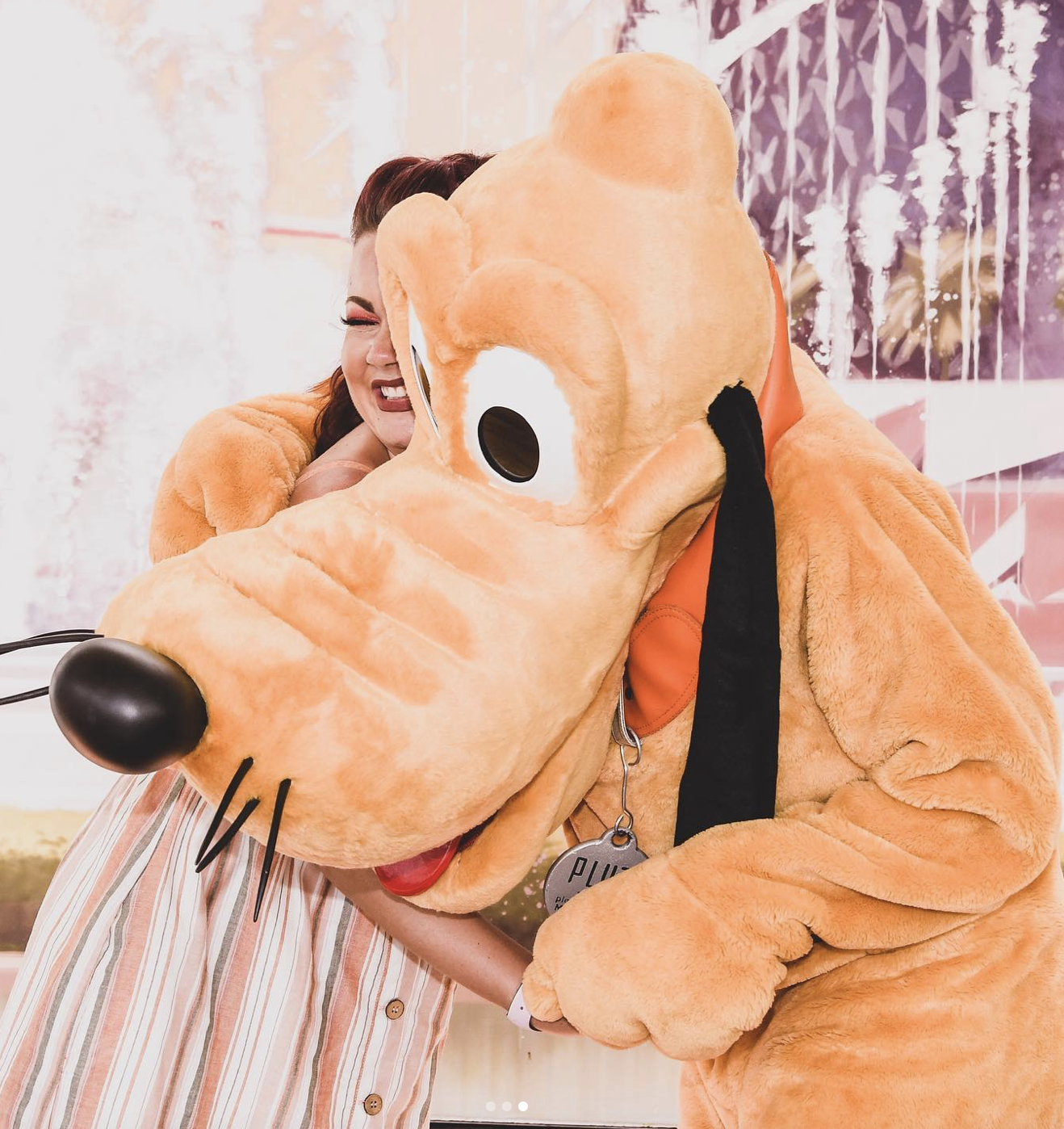 A young white woman hugs Pluto (a Disney character that is a large, yellow dog with long, thin black ears) tightly and smiles widely with her teeth, closing her eyes.