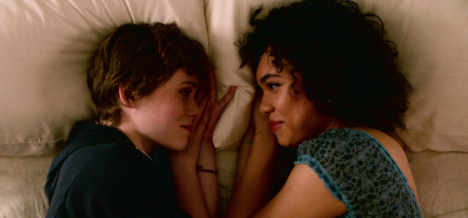 Two characters from "I'm Not Okay with This," Sydney and Dina, laying in bed facing each other. 