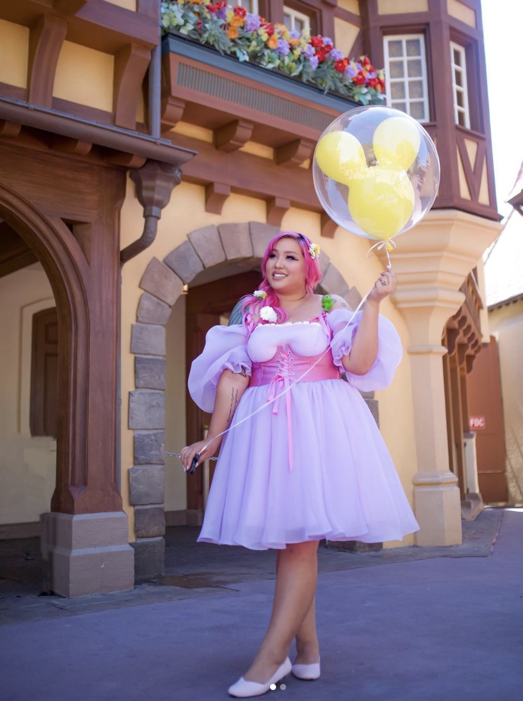 A light-skinned woman with long pink hair poses in a snort, puffy Rapunzel dress. She is holding a yellow Mickey Ears balloon and smiling off into the distance.