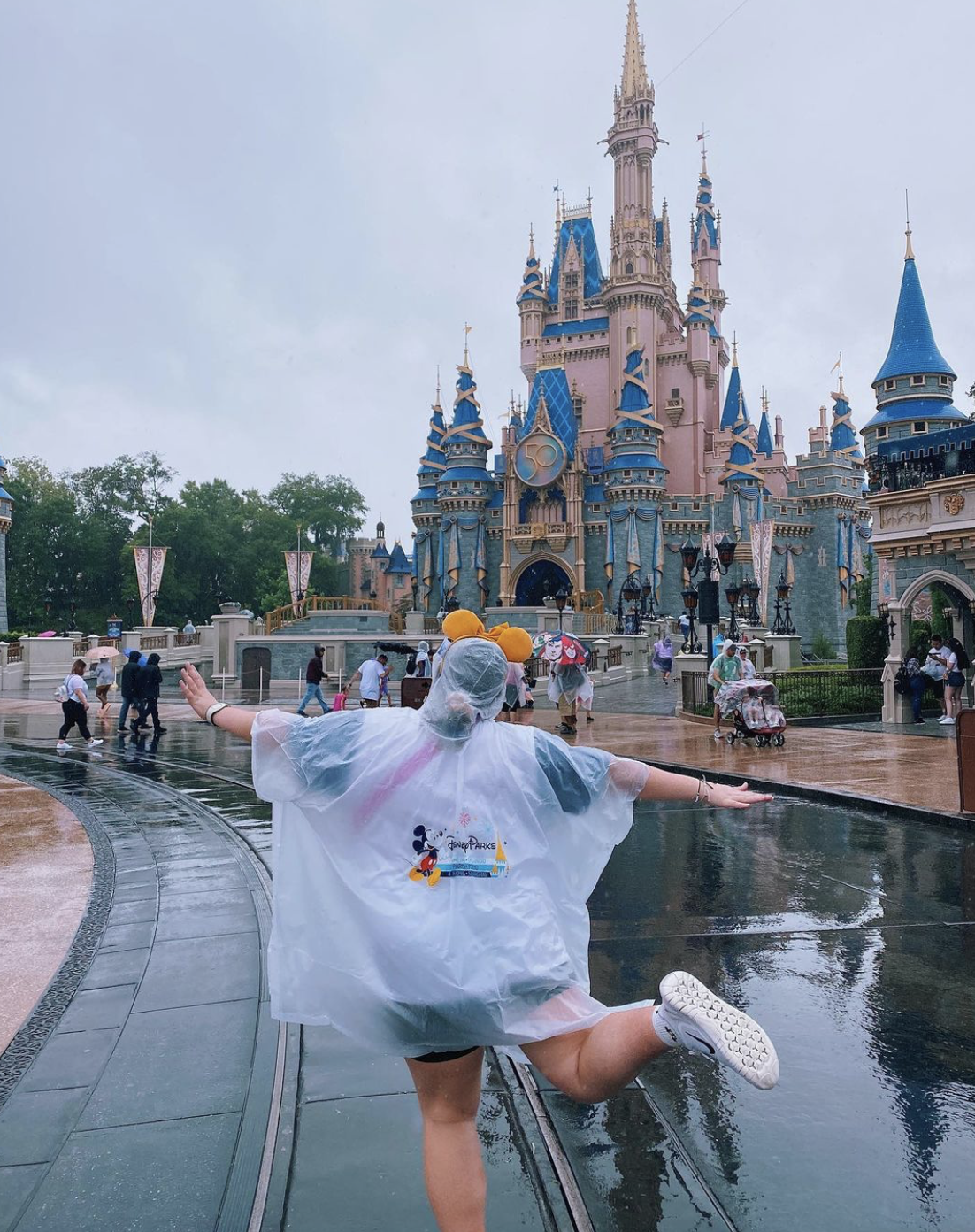A light-skinned person runs forward, arms flung out to their sides, with one leg kicked back. They are wearing a clear rain poncho with the Disney World logo on the back and using a pair of yellow Mickey Mouse ears to hold the hood to their head. In front of them, the street leading to Cinderella's Castle is glistening in the rain.