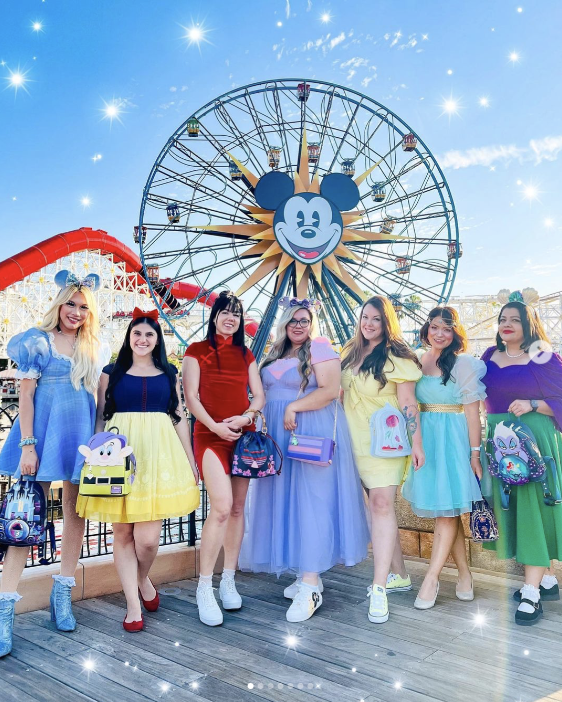 Seven light-skinned women pose in front of the Mikey Mouse ferris wheel at Disneyland. They are each dressed in clothes that resemble different Disney characters. From left to right: Cinderella, Snow White, Mulan, Rapunzel, Belle, Jasmine, and Ursula.