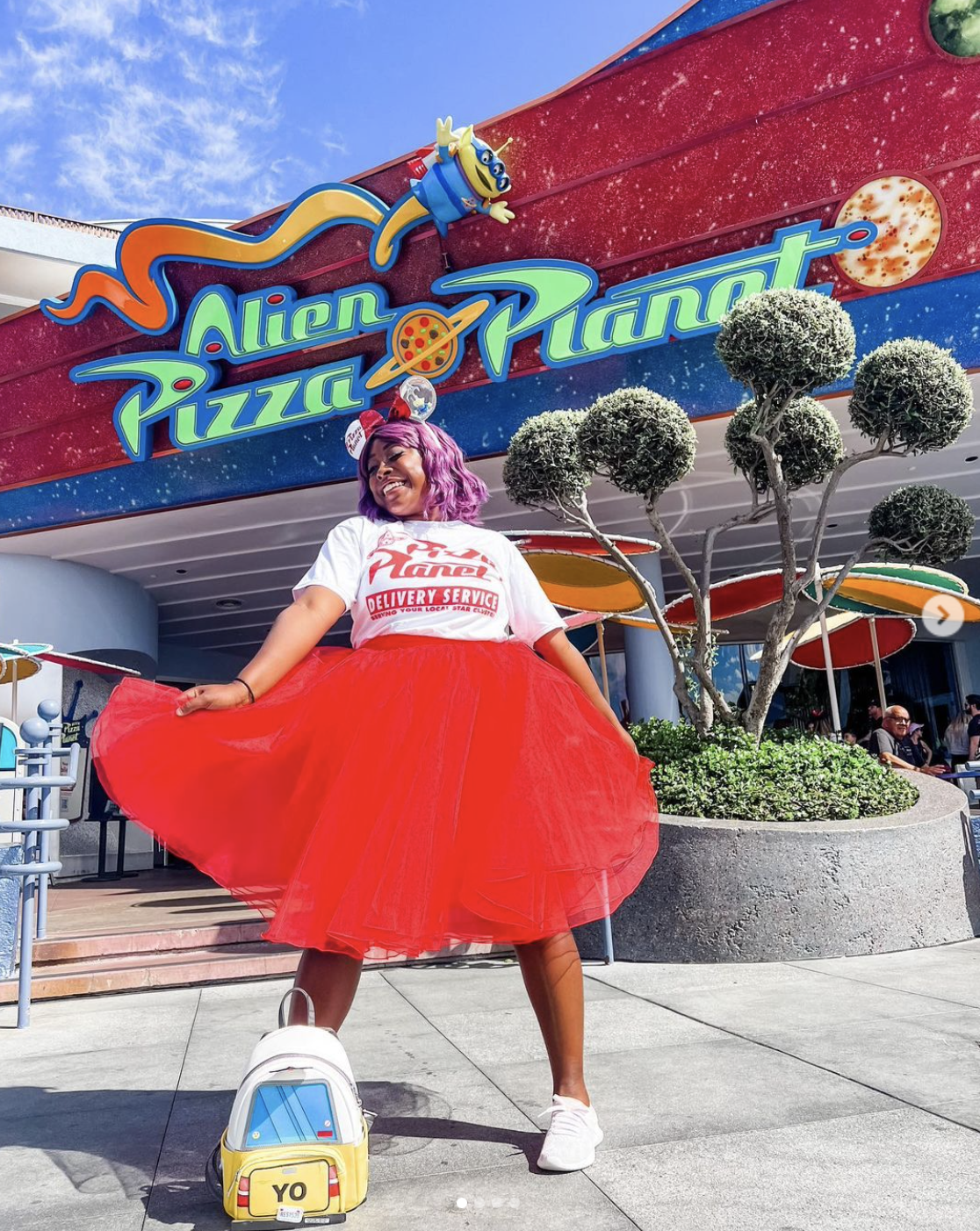 A Black woman with pink hair poses in front of the Alien Pizza Planet at a Disney theme park. She is holding her bright red midi skirt in a swishing motion as she grins.