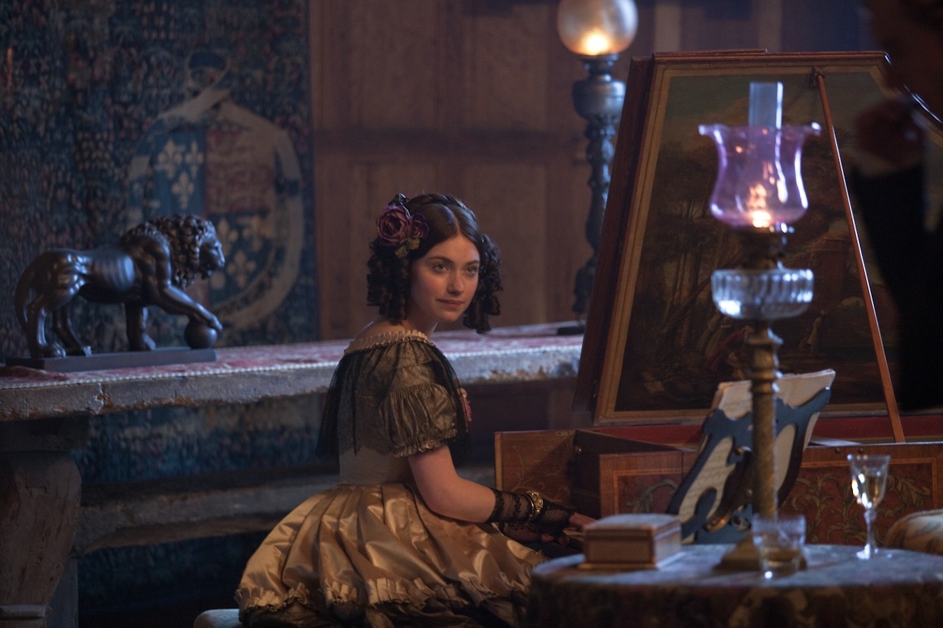 A thin white woman dressed in an elaborate period wear sits at an intricately carved and painted piano. Her hair is in tight coils, and she looks sternly past the camera. The room she is in is lit by waning daylight and two lamps. Behind her, the statue of a lion stands on a wooden table, positioned against a wood-paneled wall. A faded tapestry hangs, cluttering the shot with even more background color, pattern, and texture.