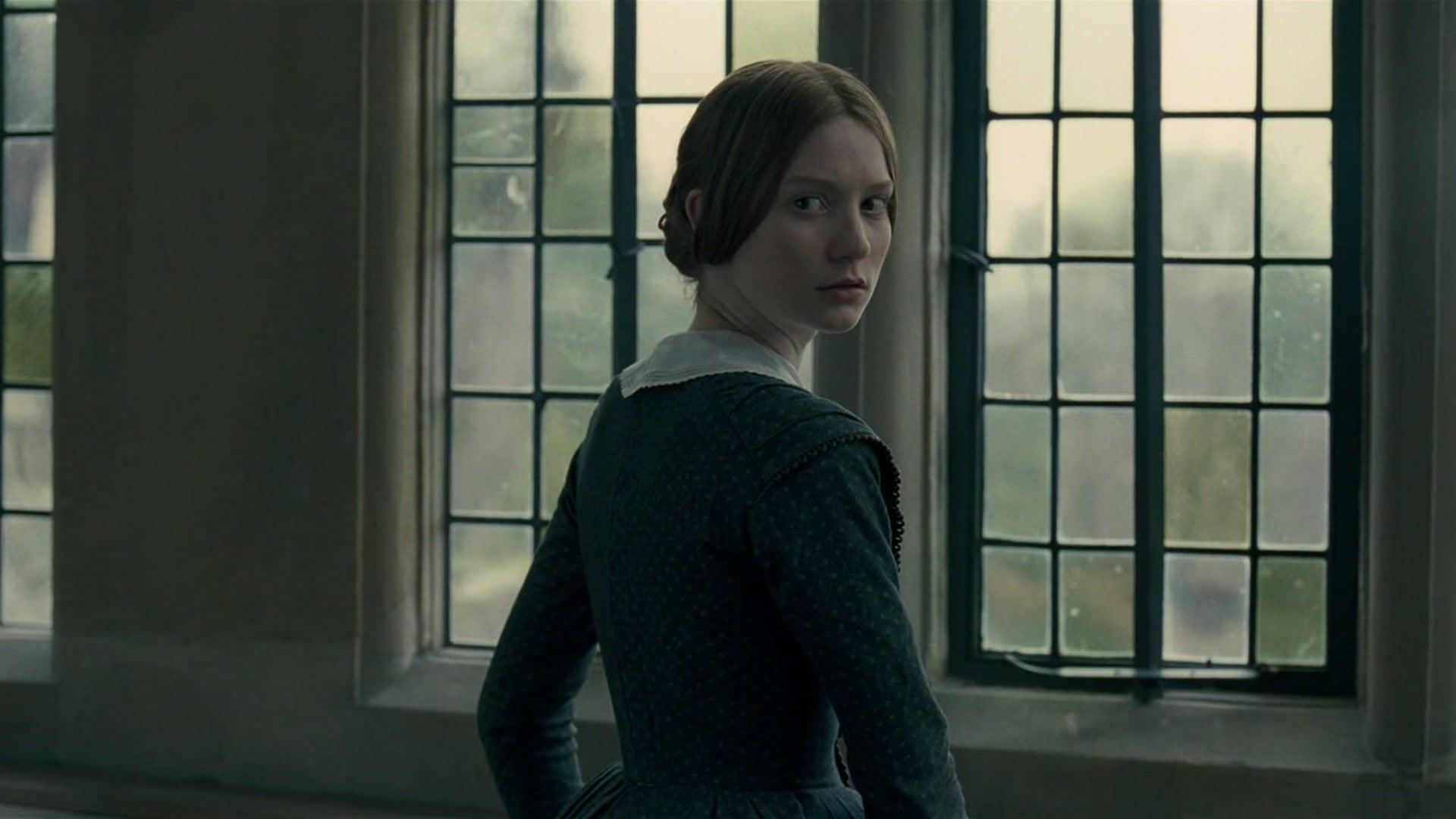 A white woman dressed in period clothing stands in front of a set of windows. She looks over her shoulder, past the camera, with a troubled expression.