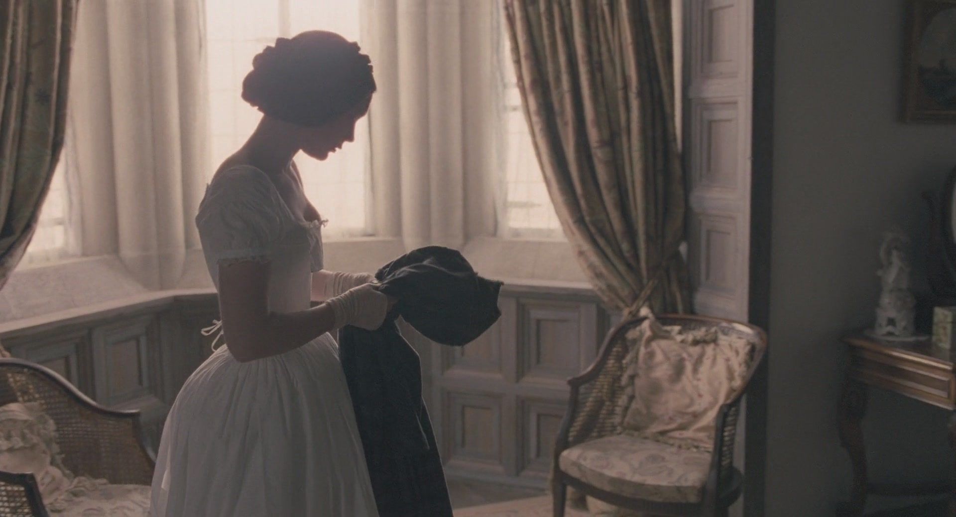 A thin, white woman stands alone in a room. Both she and the room are dressed in period style. Her hair is braided elabrately on her head and she looks down at a coat she is holding with both hands. A large set of cutoned windows filters sunlight behind her making the room look hazy.