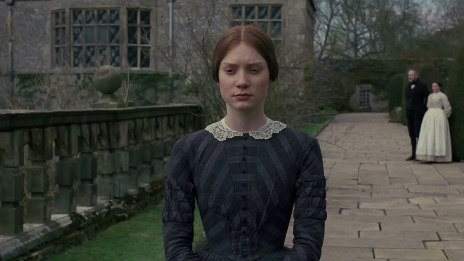 Jane Eyre: A white woman dressed in period clothing stands in front of a set of windows. She looks over her shoulder, past the camera, with a troubled expression.