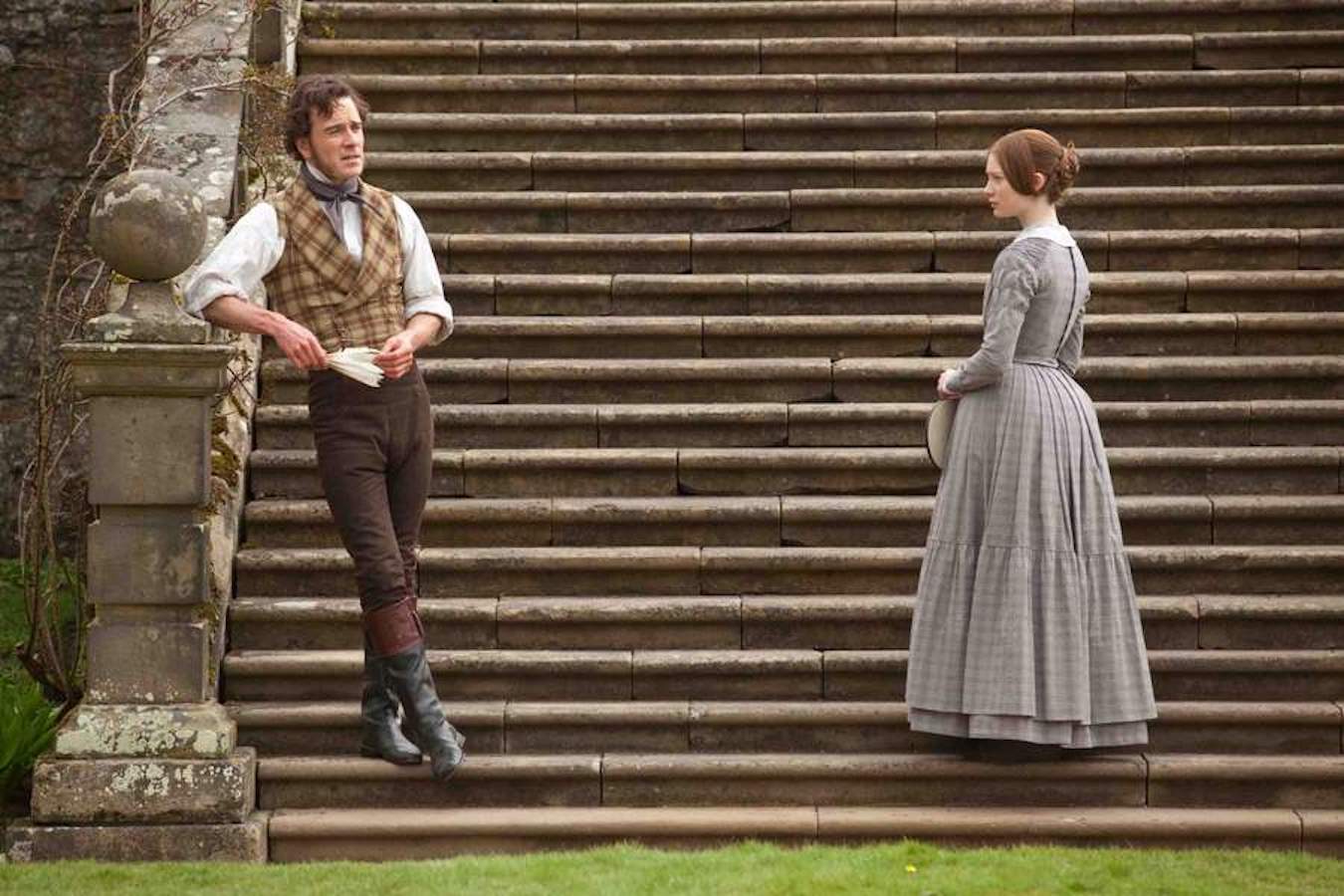 A white man and woman dressed in period clothing stand on an expansive stone staircase. They appear to be engaging in conversation although they are distanced. The man rests his elbow on the large stone railing and the woman holds her hat in both hands at her waist.