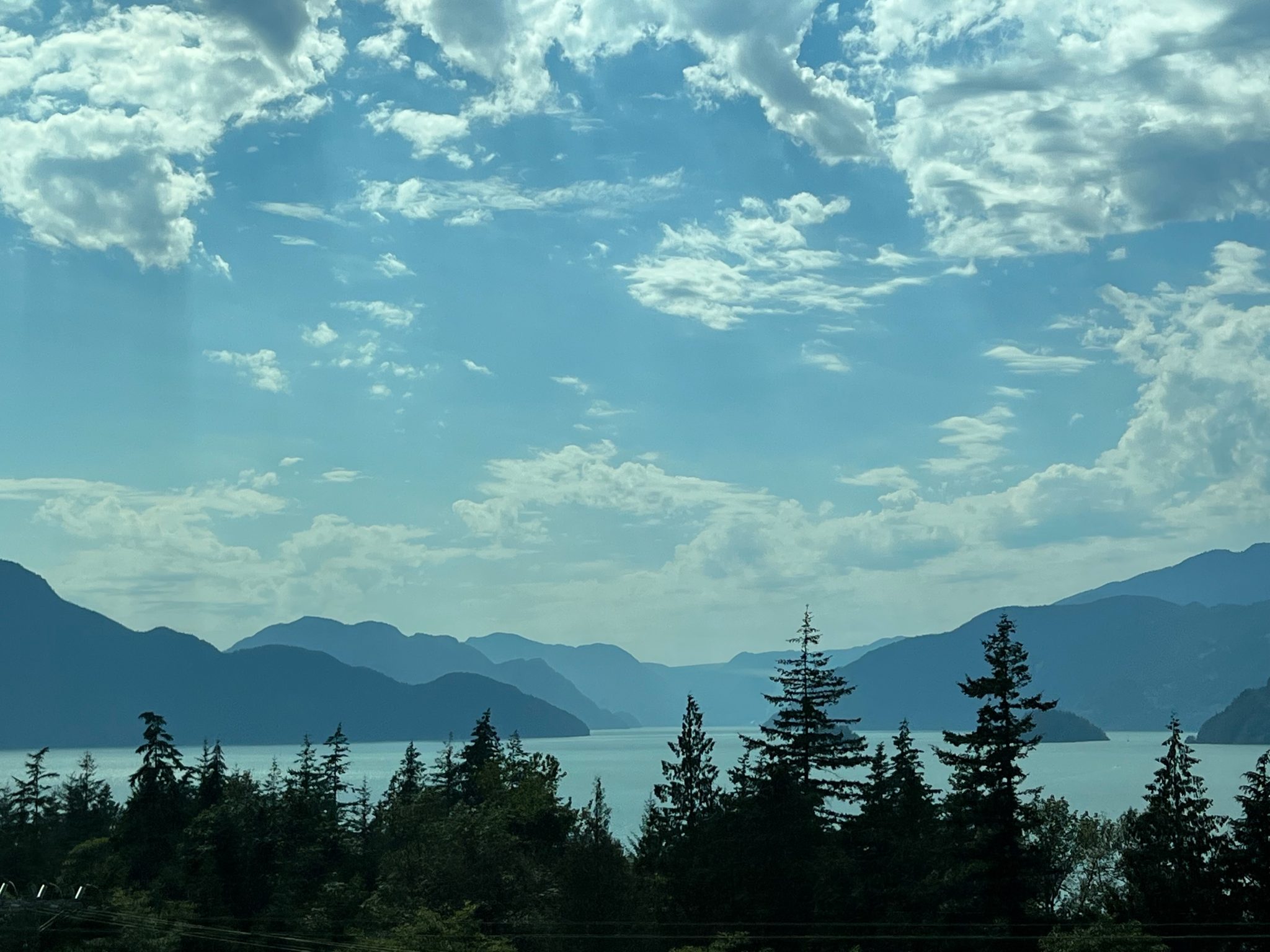 In the foreground, the dark silhouette of evergreen trees slightly blocks the view of a large lake surrounded by distant mountains. The sky is bright blue with scattered white clouds.