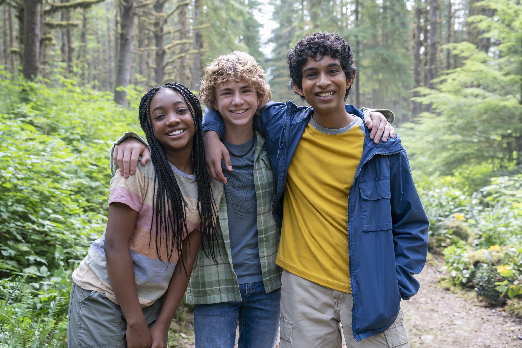 Annabeth (a young Black girl), Percy (a young white boy), and Grover (a young boy with brown skin) stand on a wooded path with their arms thrown over one another's shoulders.
