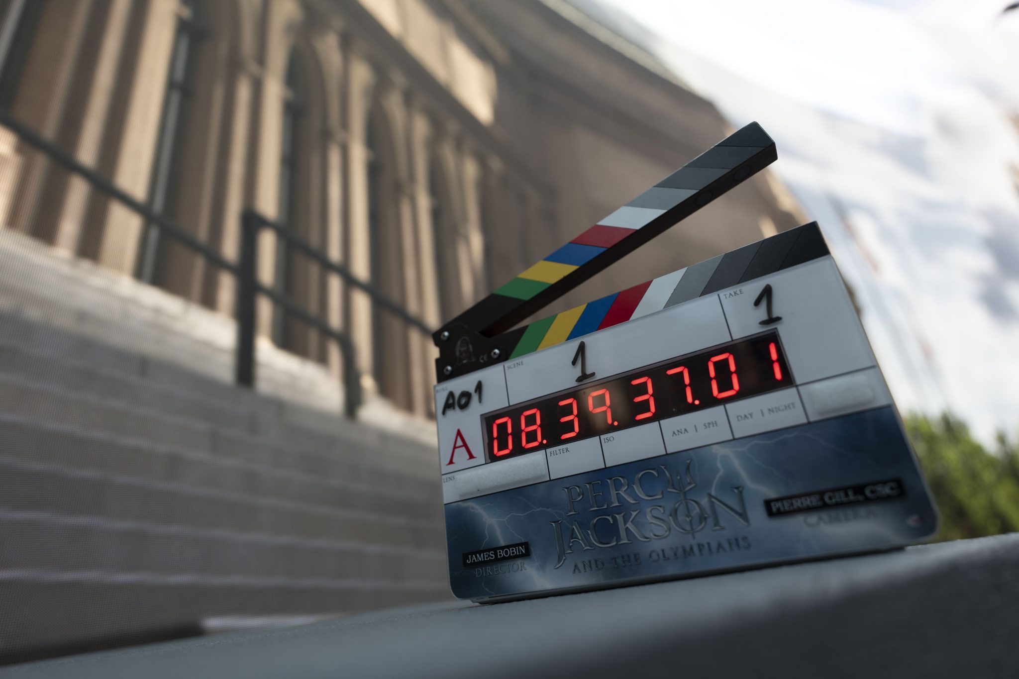 The camera sits on the ground, looking up at a set marker. In the background, a large traditional-style building looms.