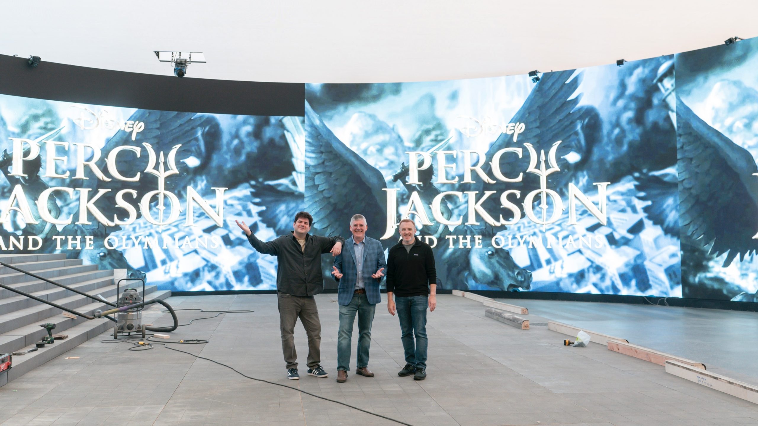 From left to right, Dan Shotz, Rick Riordan, and Jon Steinberg stand on a sound stage. Behind them, large screens show a portion of the cover of the first Percy Jackson book.