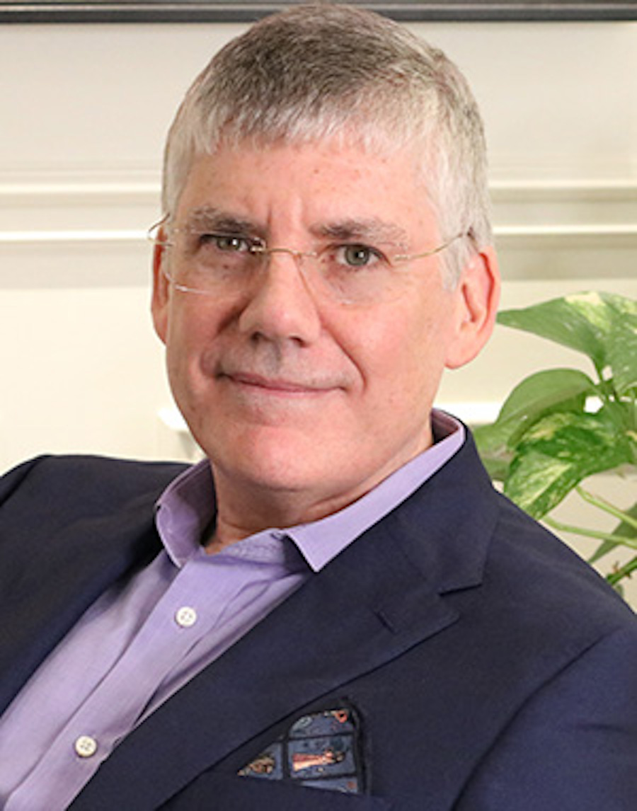 Author Rick Riordan smiles at the camera with closed lips. He is a midd-aged white man with grey hair, thick eyebrows, and wire-rimmed rectangle glasses. He is wearing a lavender dress shirt with a navy blue suit jacket. He looks friendly and smart.