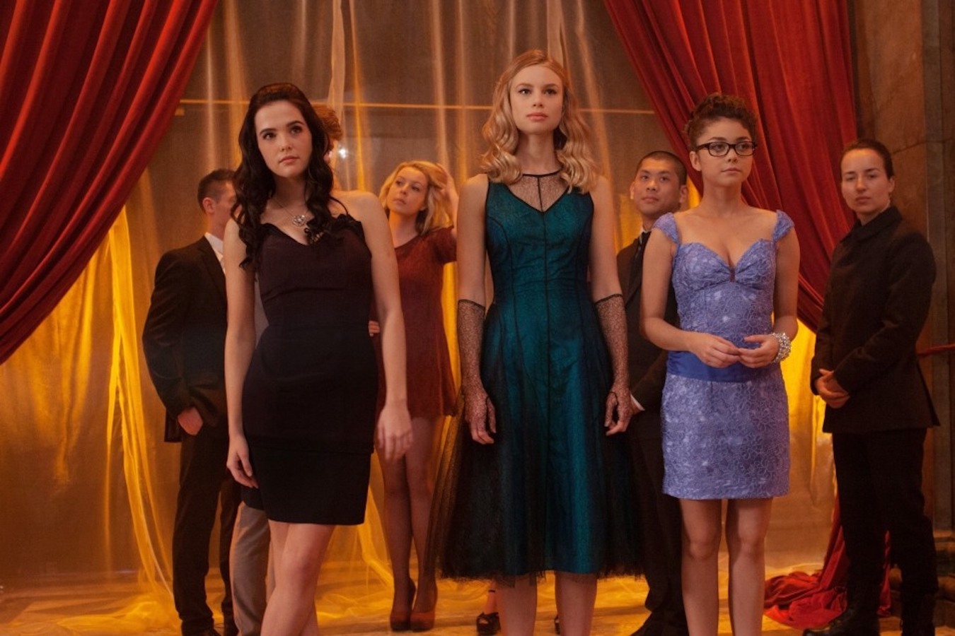 Students stand near the curtained entrance to an elegant school dance. A tall, blonde, white woman stands with her arms at her sides; she is wearing an emerald-green dress and fishnet gloves. Flanking her are two shorter, brunette women. One strikes a poised pose; the other stands awkwardly.