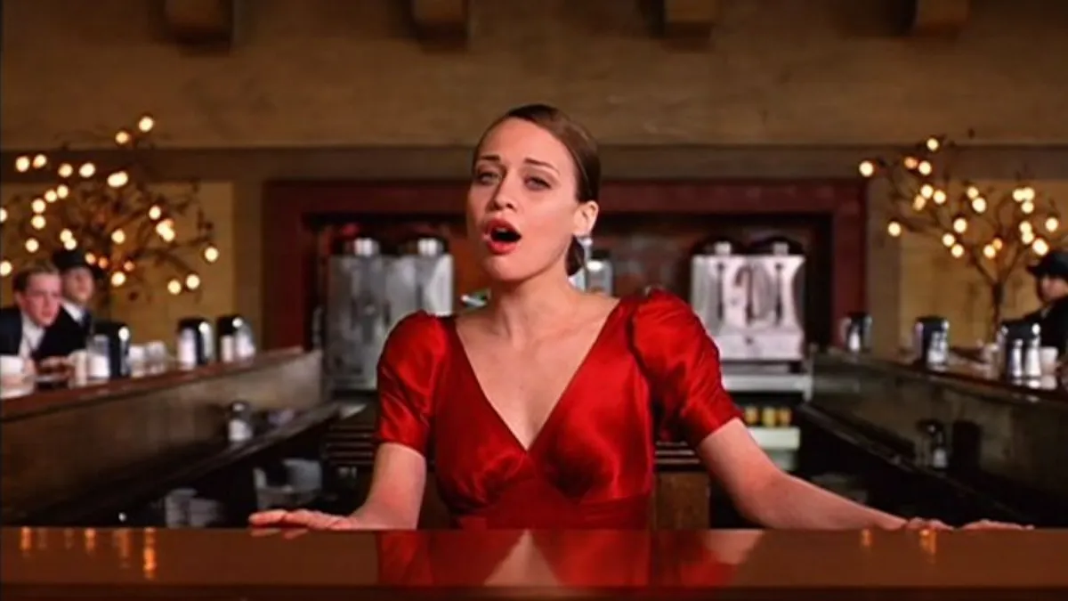 Fiona Apple dons a red, silky dress while placing her hands on the shiny (drink) bar in front of her, slightly leaning back in her music video for the song, "Paper Bag."