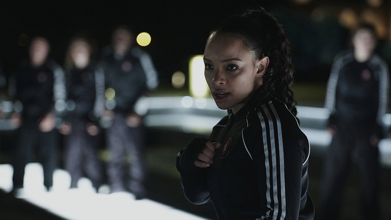 A young Black woman stands, hands held in a fighting stance. Her hair is pulled into a ponytail. She is staring down her training opponent somewhere behind the camera. Behind her, the other students watch.