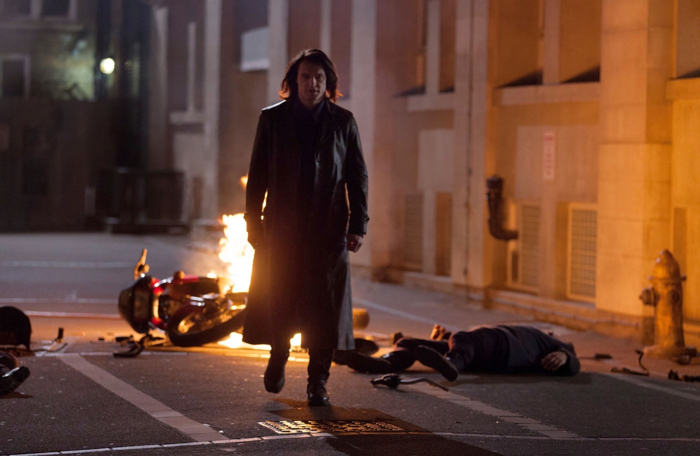 A tall man in a long, black trench coat strides purposefully toward the camera. Behind him, a motorcycle lays in flames on the city street, and a person lays incapacitated on the ground. 