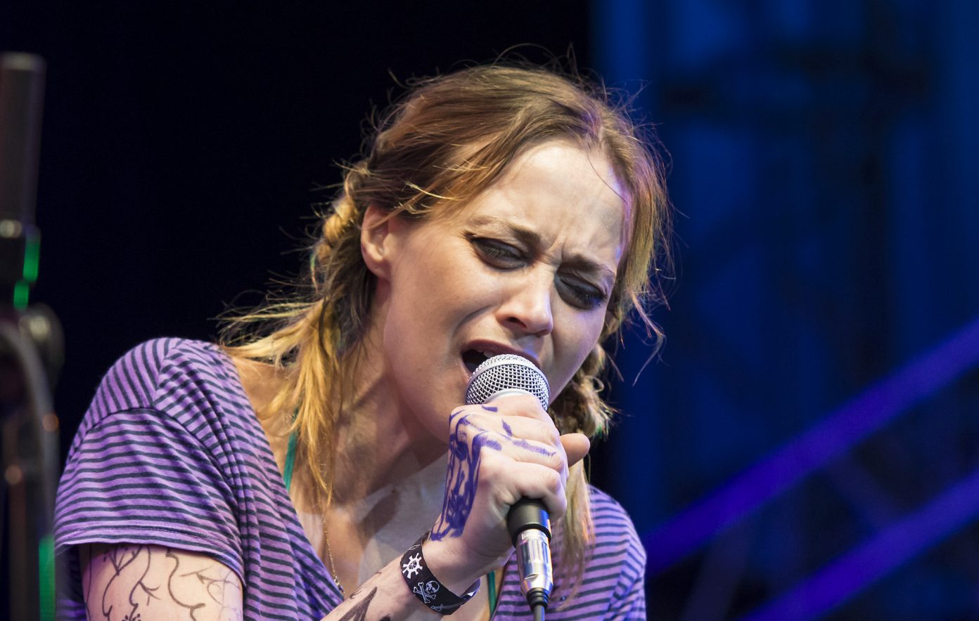 Fiona Apple sings hunches over slightly as she sings passionately into a microphone, her brown hair with sandy blond highlights in messy, short braids.