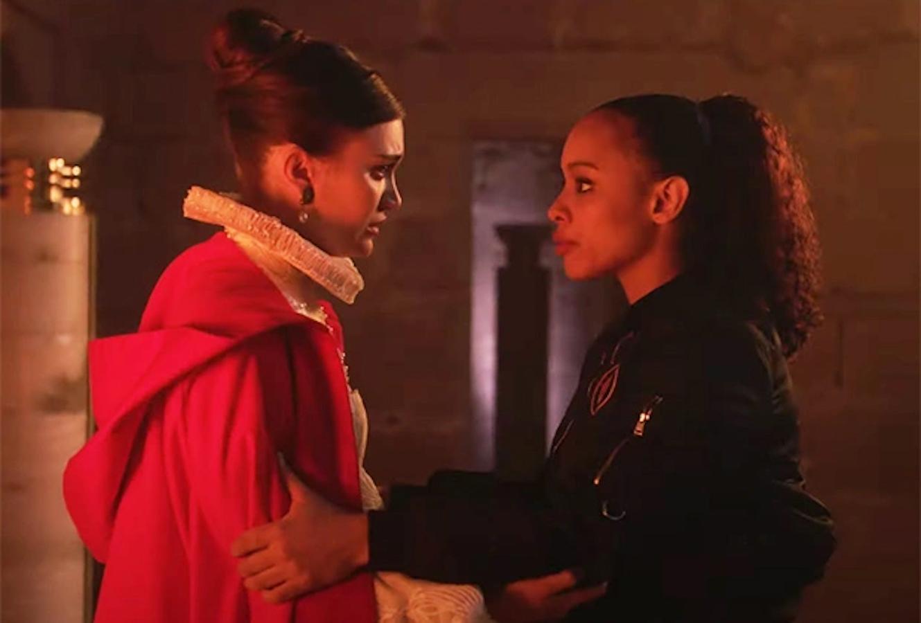 A young, brunette, white woman dressed in elaborate, collared robes looks anxiously into the eyes of her friend. A young Black woman stands opposite the first; she is dressed much more informally. They clutch onto one another’s arms.