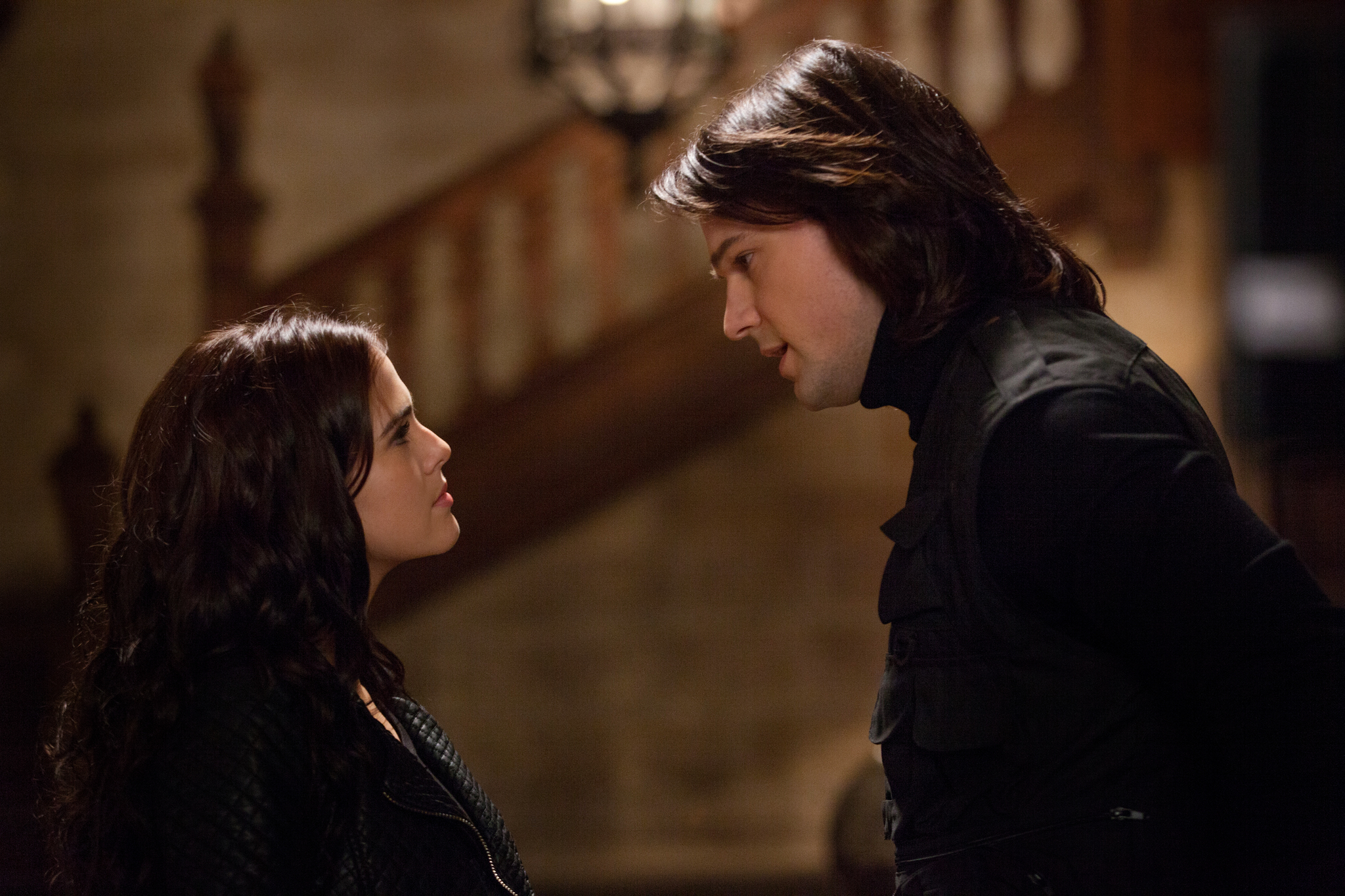 In a still from Vampire Academy (2014), a tall, white man with shoulder-length brown hair looks down as he is speaking. Facing him is a brunette white woman who is shorter than him. She has a stubborn look on her face.