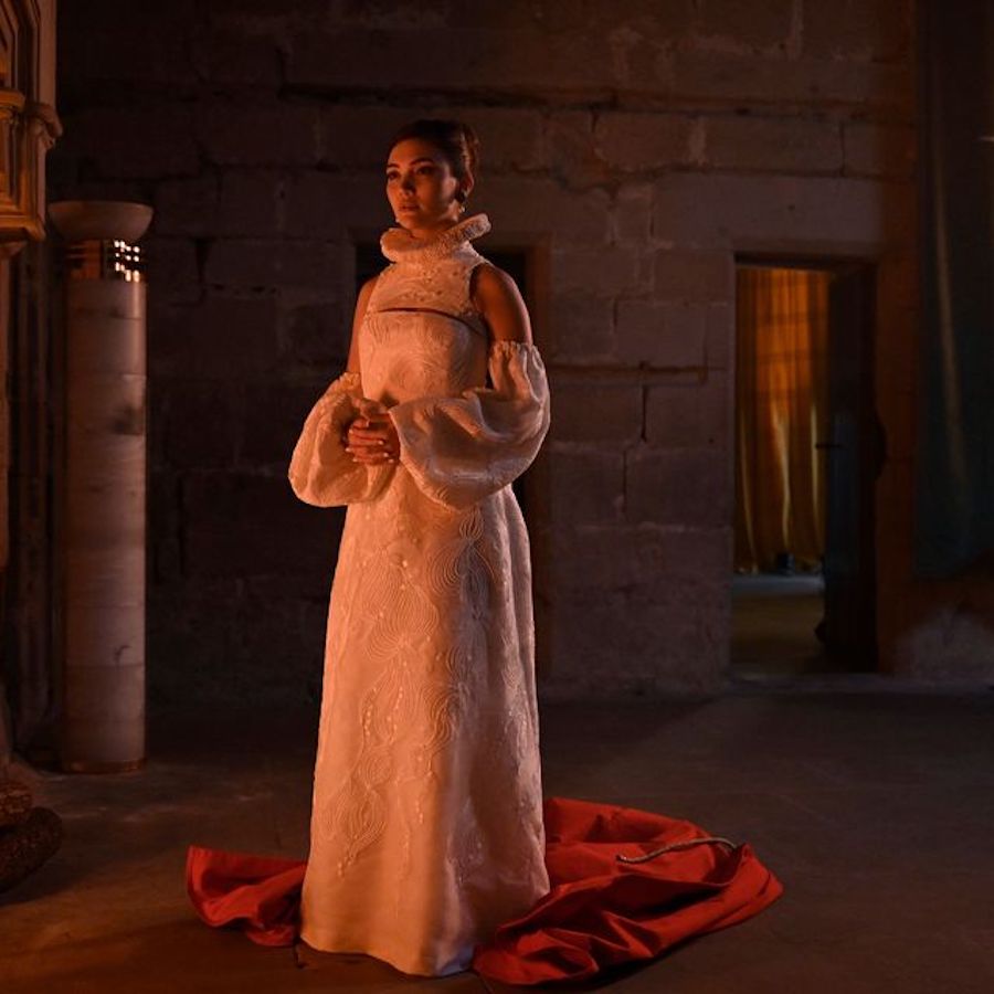 A thin, brunette, white woman stands in a barren castle room. She is wearing an elaborately beaded dress with detached sleeves and a dramatic collar. Red fabric is pooled around her feet. Her hands are clasped at her waist and she stares purposefully undaunted past the camera.