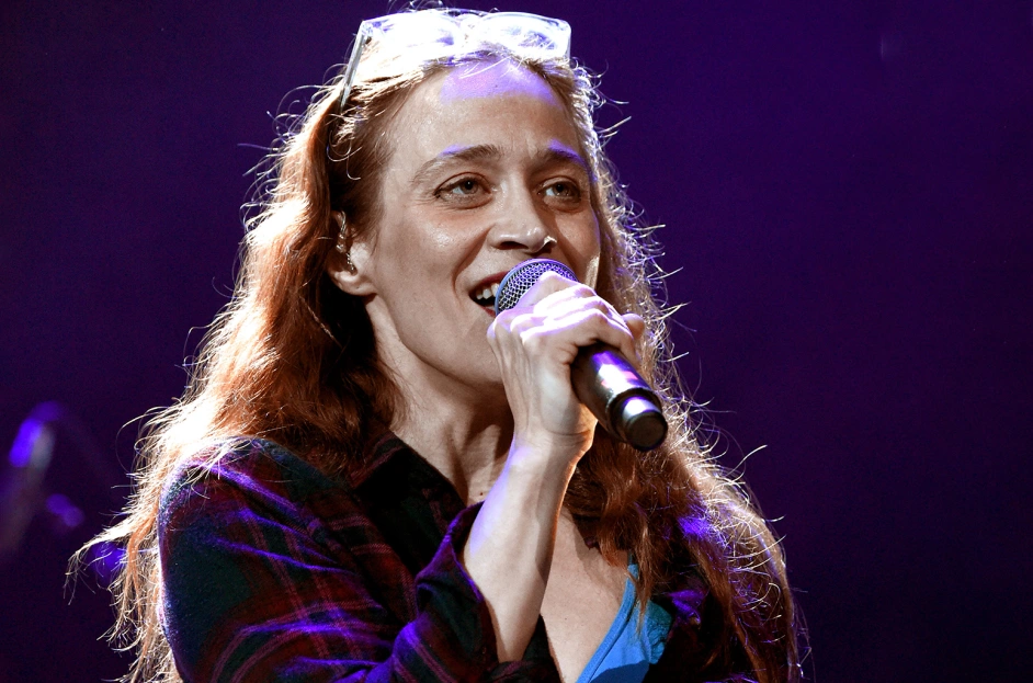 Fiona Apple smiles as she sings into a microphone, her face illuminated by a spotlight.