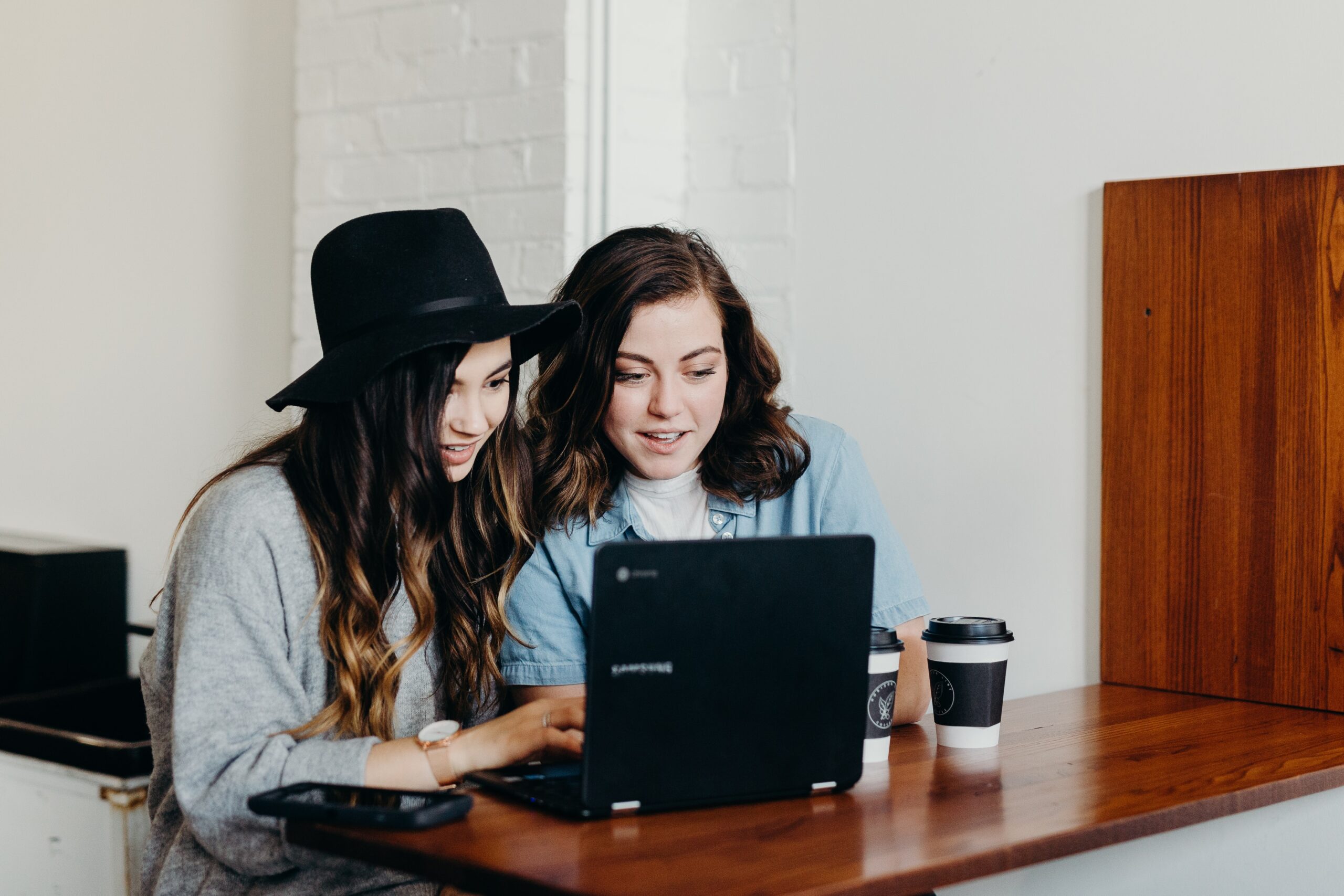 Two women sitting in front of a laptop smiling and drinking coffee. 