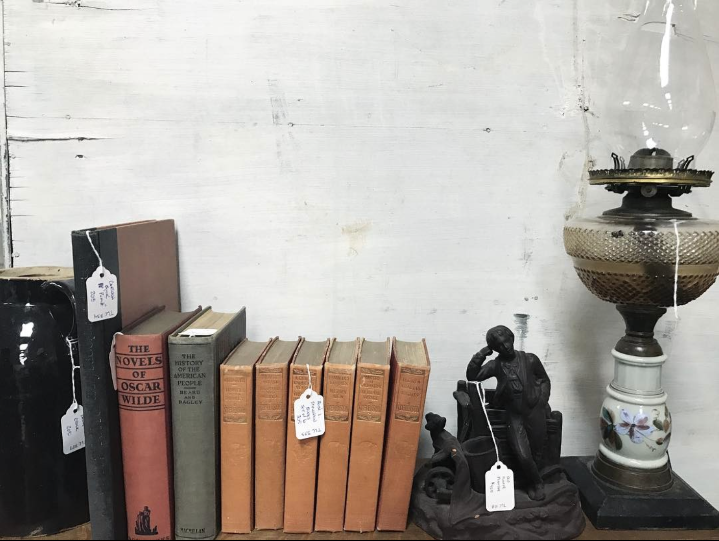 A set of antique books sits on a shelf next to other antique items. Small white price tags hang from some of them, indicating that they are for sale.