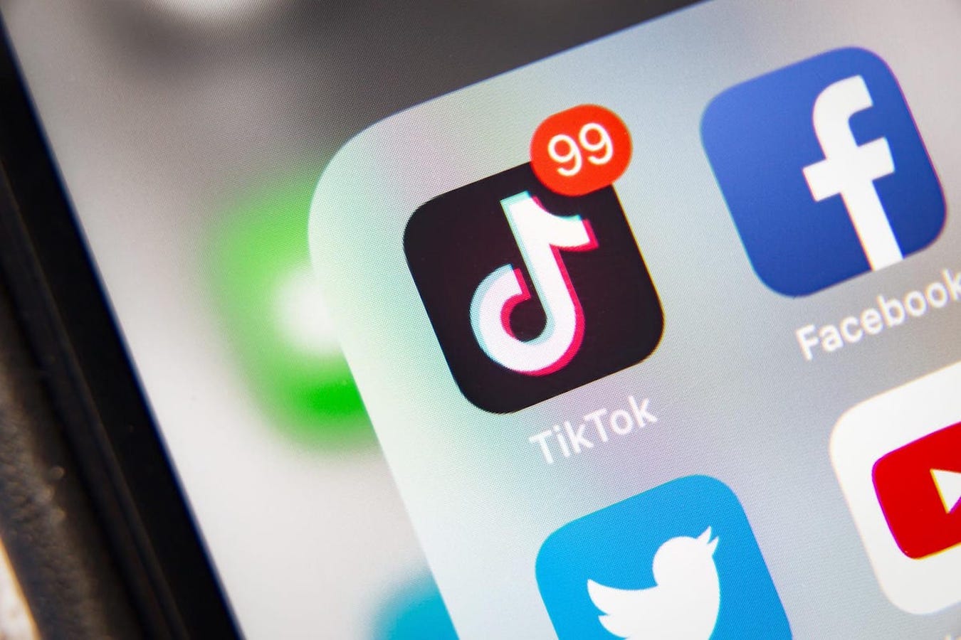 A close-up of an app folder on an iPhone shows the TikTok app next to Facebook, Twitter, and YouTube. There are 99 unread notifications for TikTok.