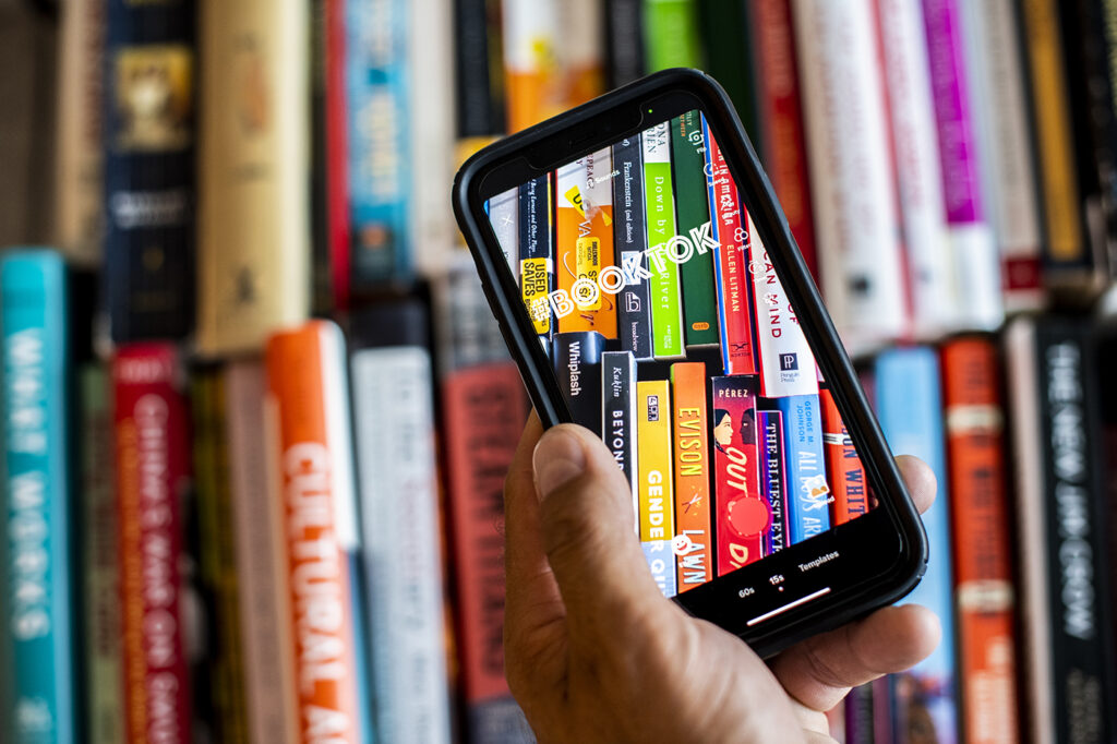 A hand is holding a cell phone. They are taking a photo of several book spines lined up.