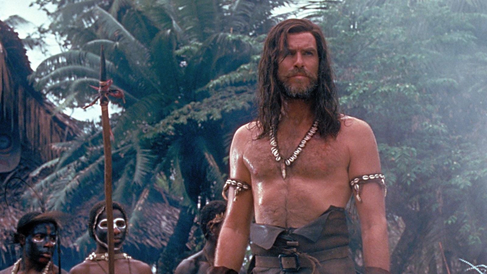 A film still from the 1997 adaptation of Robinson Crusoe. Crusoe, a white man, stands shirtless, looking forward at something off camera. He is scruffy but mostly well-kept. Behind him, several dark-skinned Black men depicted with face paint and holding spears, look on.