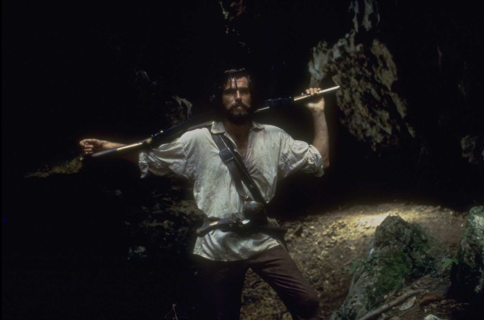 A film still from the 1997 adaptation of Robinson Crusoe. Crusoe, a white man, stands in what appears to be a dark cave, holding some type of weapon over his shoulders. He is still clothed in the clothes he washed ashore in.