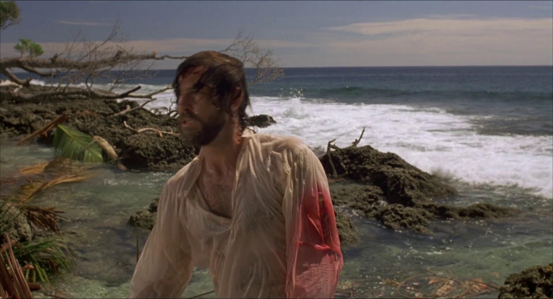A film still from the 1997 adaptation of Robinson Crusoe shows Crusoe freshly washed ashore after the shipwreck. He is standing on a debris-strewn beach, his white shirt-arm stained with blood.