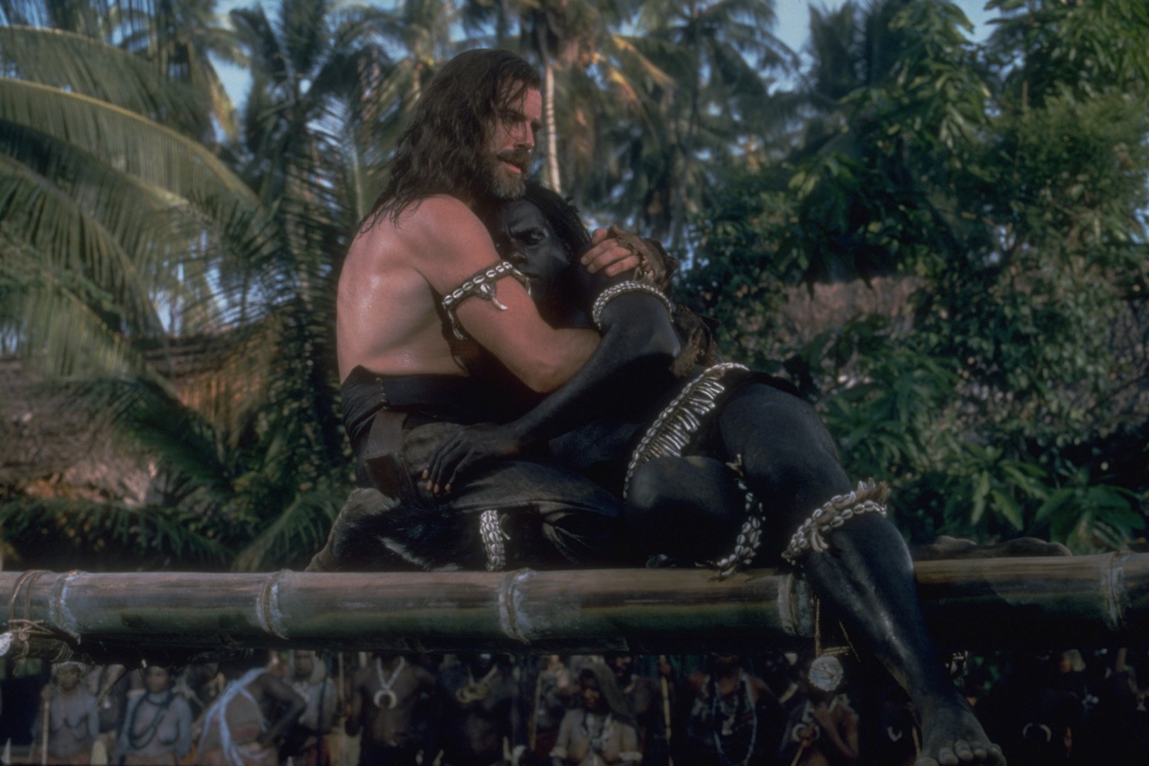 A film still from the 1997 adaptation of Robinson Crusoe. Crusoe, a white man, cradles Friday, a dark-skinned Black man after rescuing him from cannibals.