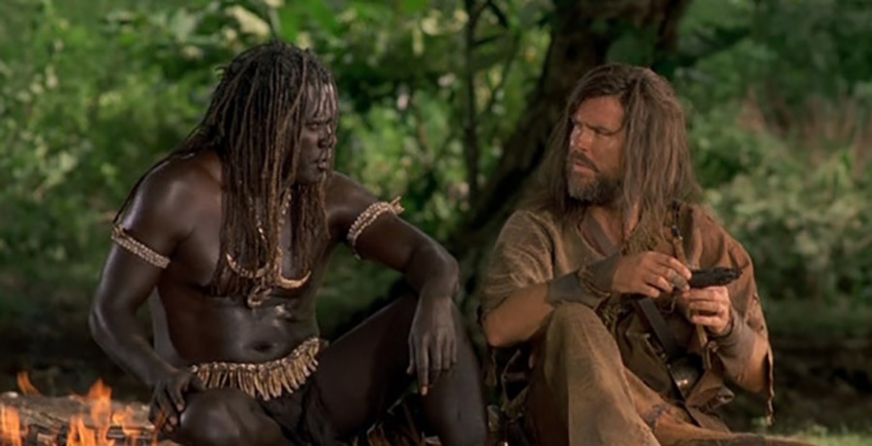 A film still from the 1997 adaptation of Robinson Crusoe. A white man, Crusoe, and a dark-skinned Black man, Friday, sit on the ground next to one another with their heads turned to face each other in conversation. Both men have long hair, but the Black man is wearing only a loin cloth while the white man is fully dressed.