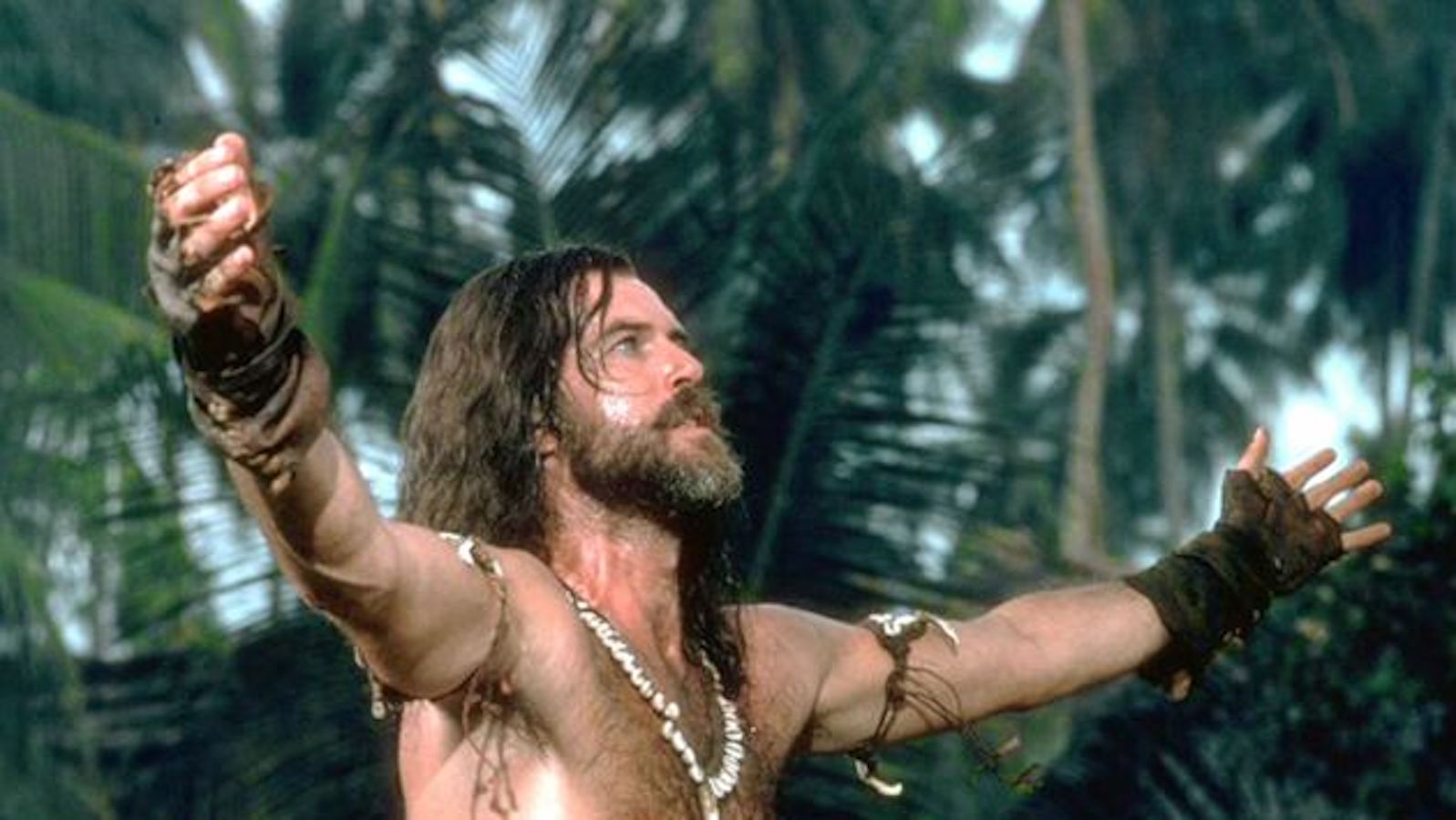 Pierce Brosnan in character as castaway Robinson Crusoe. He is a white man with long hair and an unkept beard. He is standing facing the right, shirtless, arms open wide, and looking up at something out of frame.