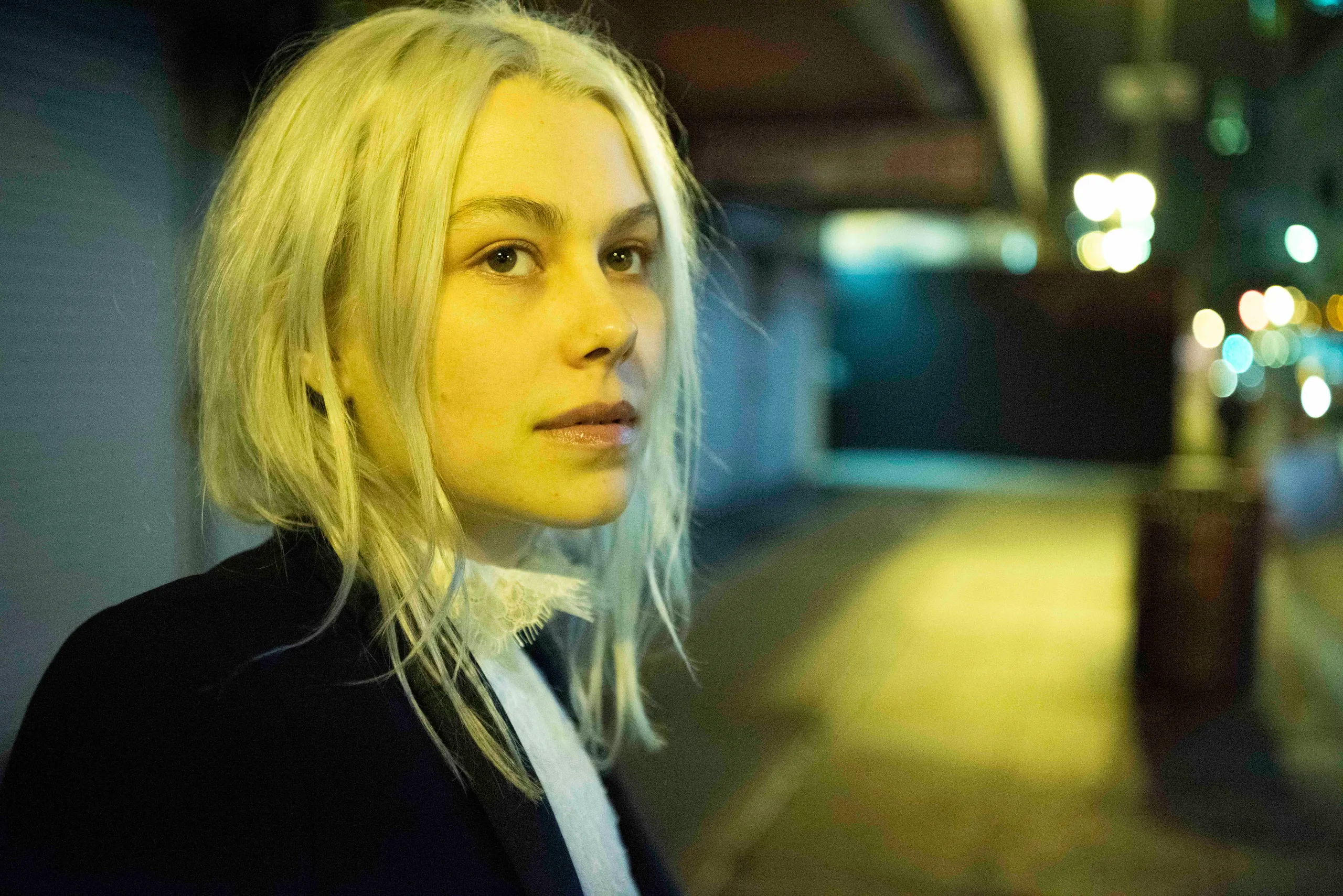 Photo of Phoebe Bridgers looking into out into the distance. She dons a black suit jacket and high-collared white shirt, her platinum hair messy, blowing in the wind. 