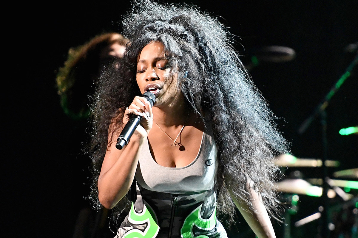 SZA sings into a mic onstage with her eyes closed. The spotlight illuminates her and her hair blows in the wind. 