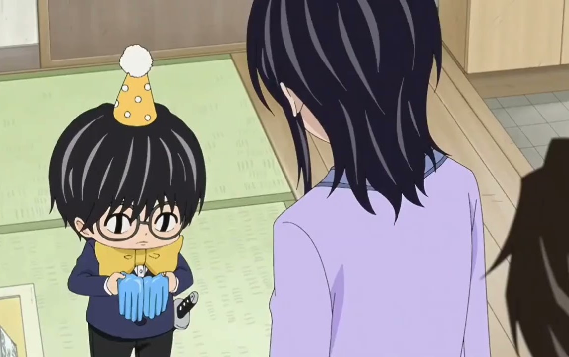 Boy wearing a yellow party hat and round glasses holds a pair of gloves for a woman to see. Episode 8. Mami Tsumura. Kotaro Lives Alone. LINDENFILMS. 2022. 