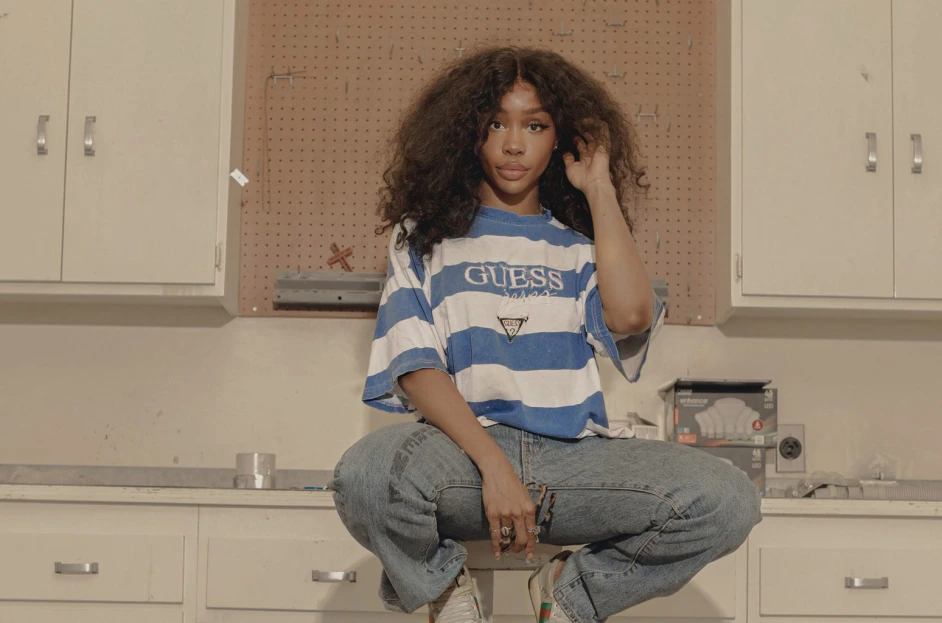 SZA sits on a stool in front of several cabinets in what looks to be a crafting/handy workspace. She tucks her hair behind her ear and looks pointedly into the camera with a very, very slight smile.