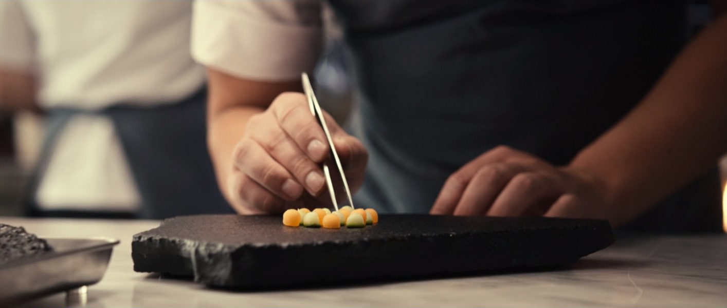 A chef is plating the amuse-bouche course of the dinner, with small yellow and orange melon balls on a black slab, in The Menu (2022).