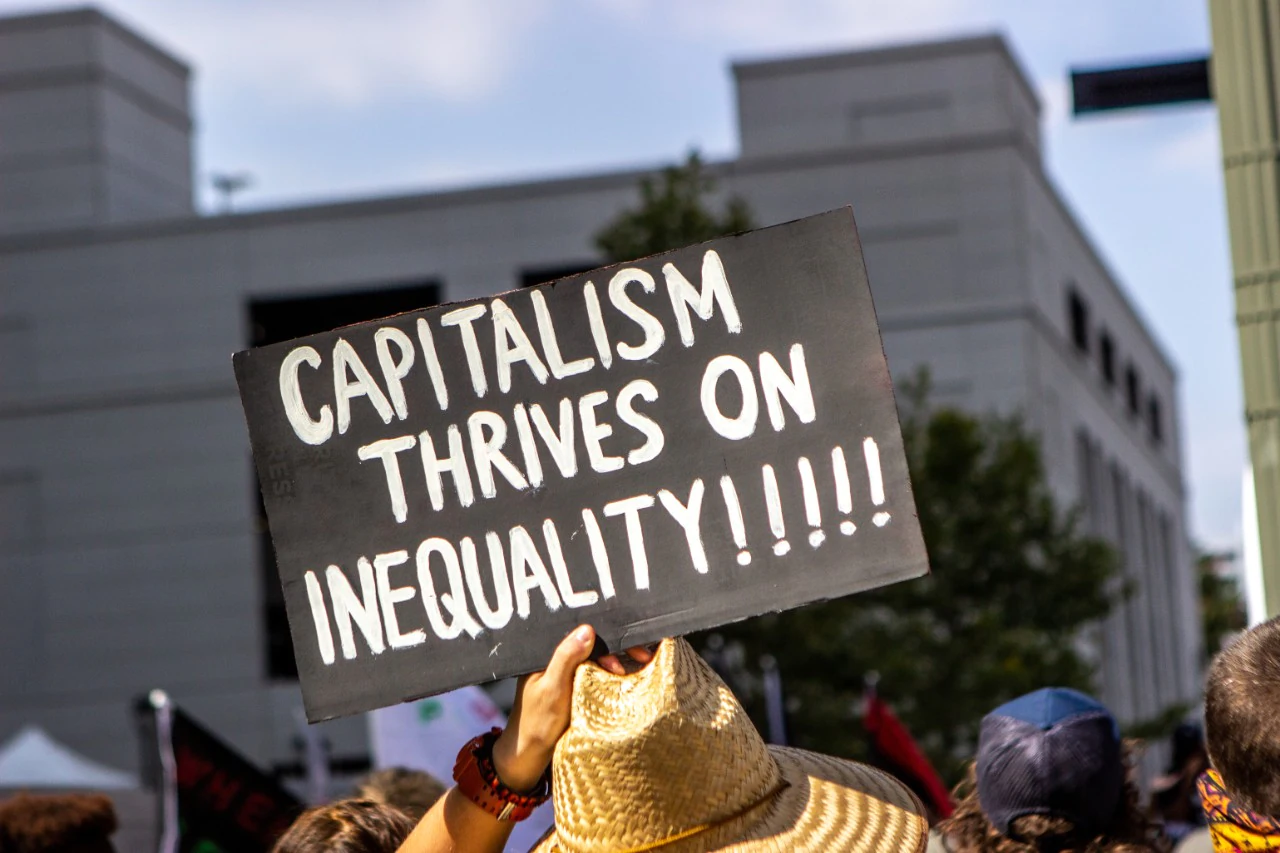 Someone holding up a sign at a protest that says, “Capitalism thrives on inequality!!!!!”