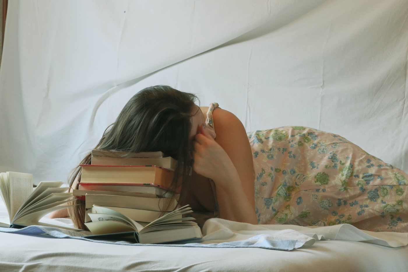 A white woman in a floral dress lays on a bed. Her head is propped up on a stack of books.