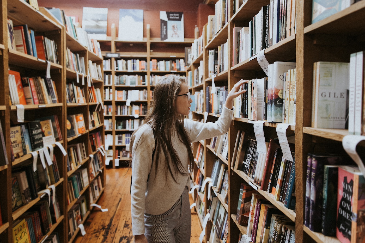 A white woman with long hair stand in between shelves of books. Her arm is extended, selecting a book from one of the shelves.