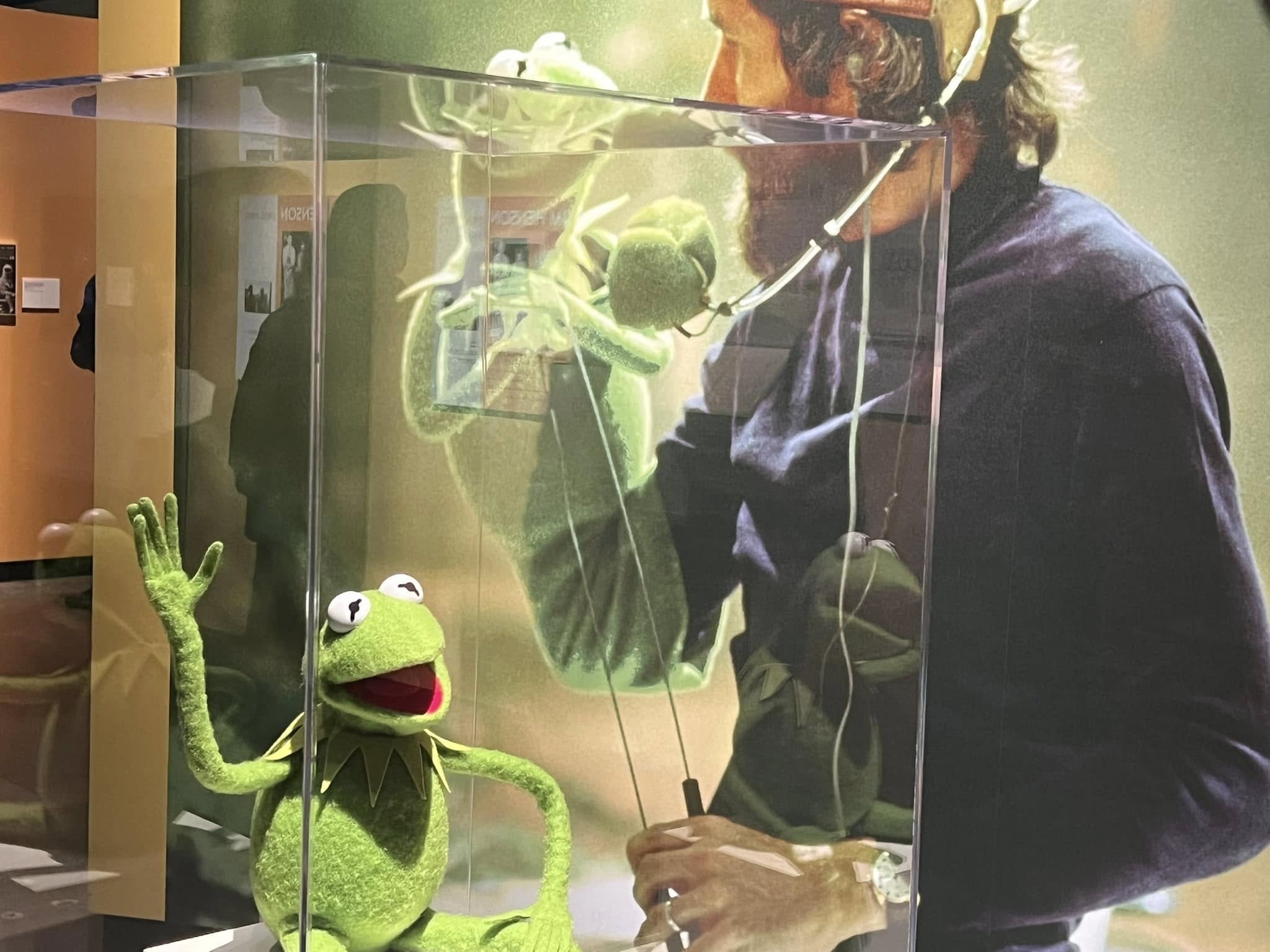 Kermit, in a glass case, with a large scale photo of Jim Henson behind him. 